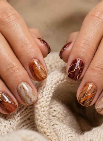 Hands with short, almond-shaped nails featuring marbled brown, beige, and gold nail art. The person wears thin gold rings and rests their hands on a textured beige fabric.