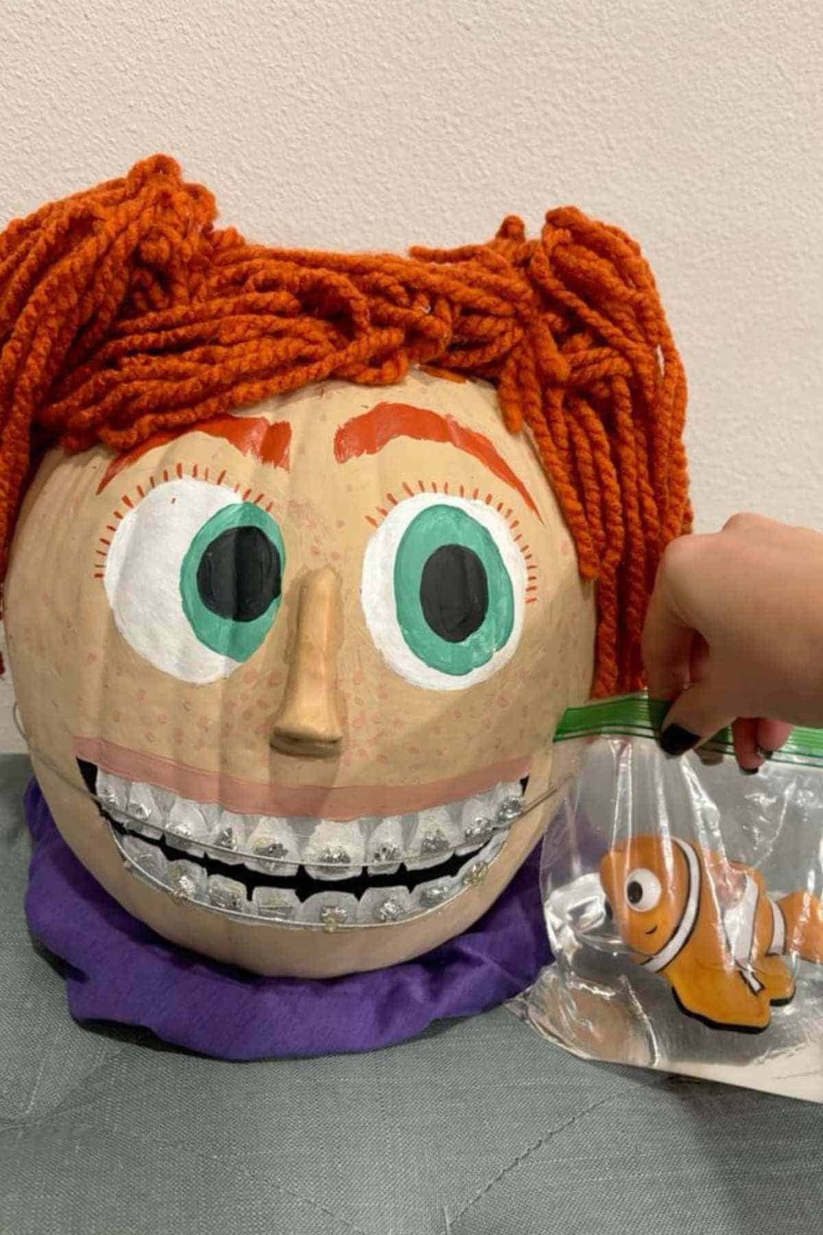 A pumpkin decorated with orange yarn hair, large painted eyes, and braces, next to a hand holding a plastic bag containing a toy clownfish.
