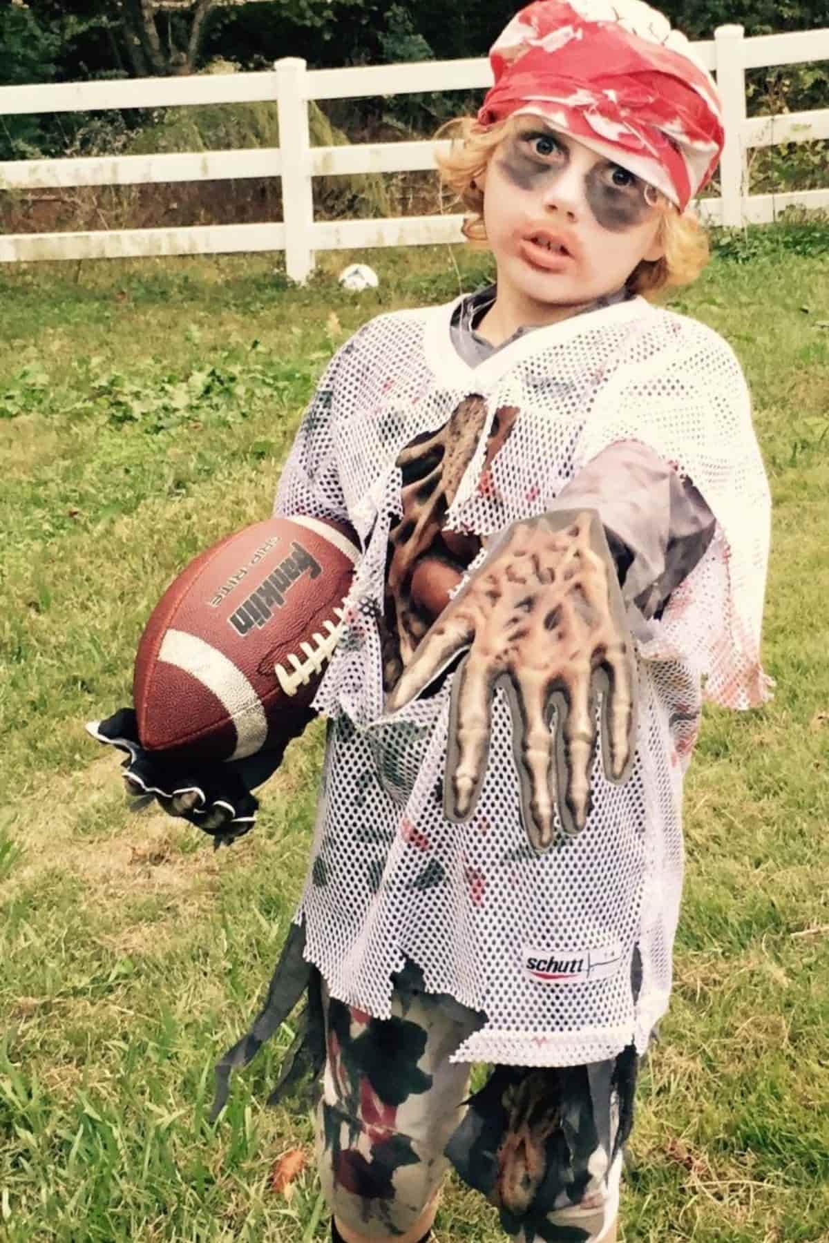 Child in a zombie football costume with face paint, tattered jersey, clawed gloves, and a football, standing on grass near a white fence.