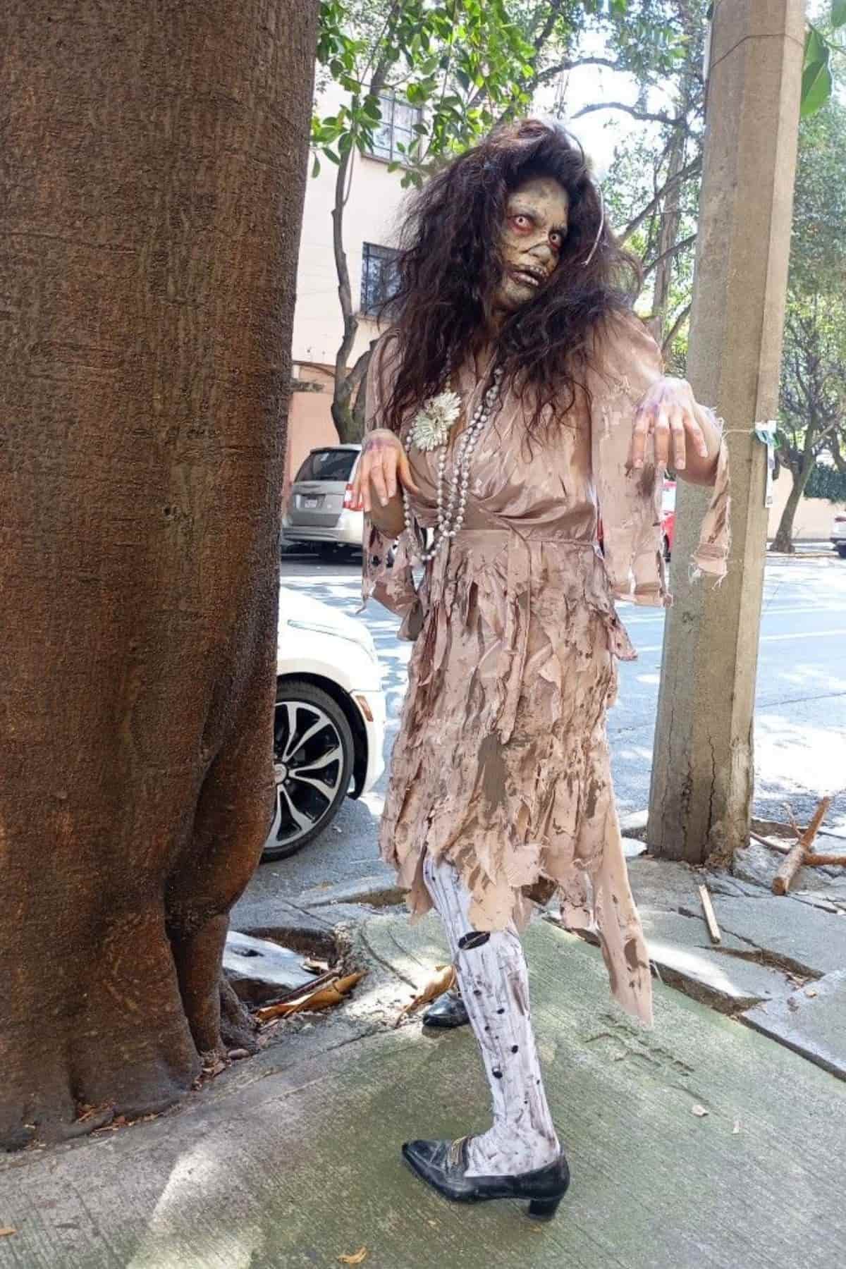 Person in tattered, beige zombie costume with messy hair, pearl necklace, and face makeup stands outdoors near a tree on a city sidewalk.