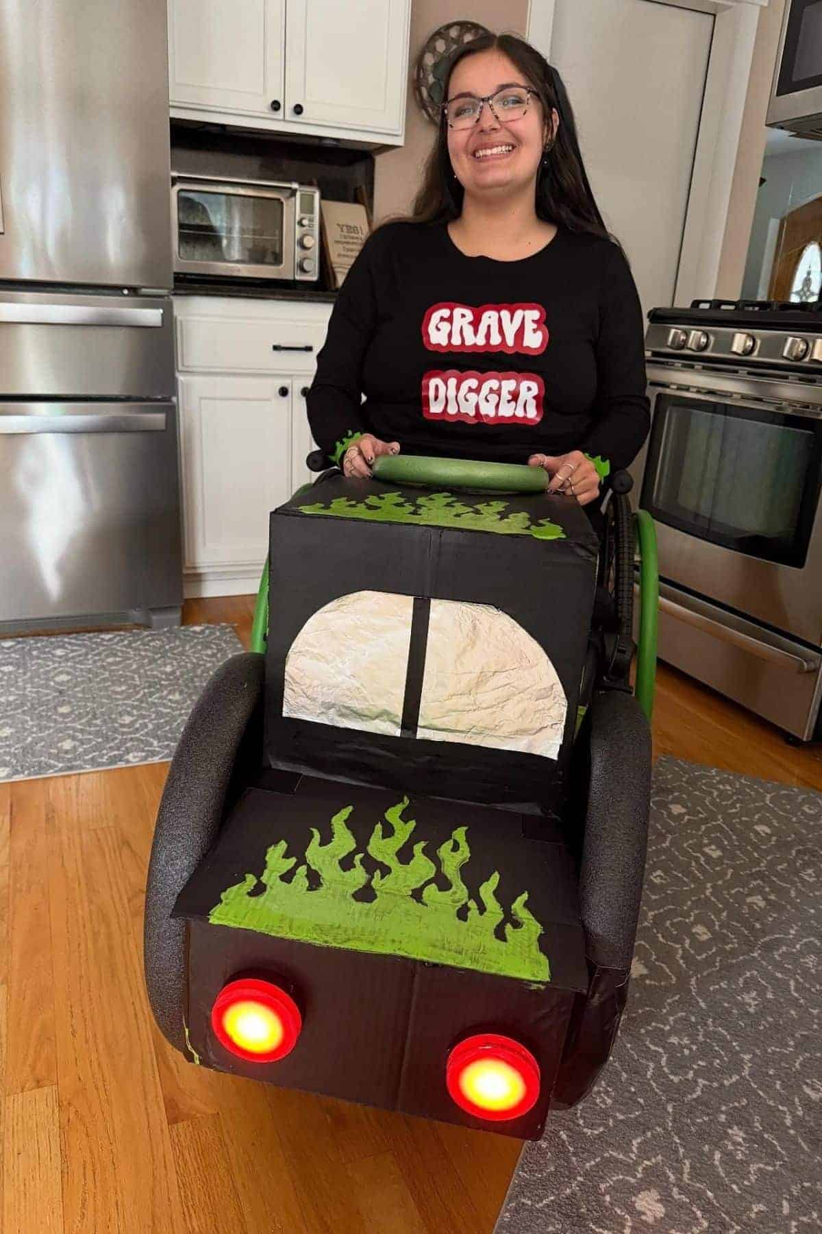 A smiling woman wearing a “Grave Digger” shirt sits in a wheelchair decorated like a monster truck with green flames and red tail lights, in a kitchen.