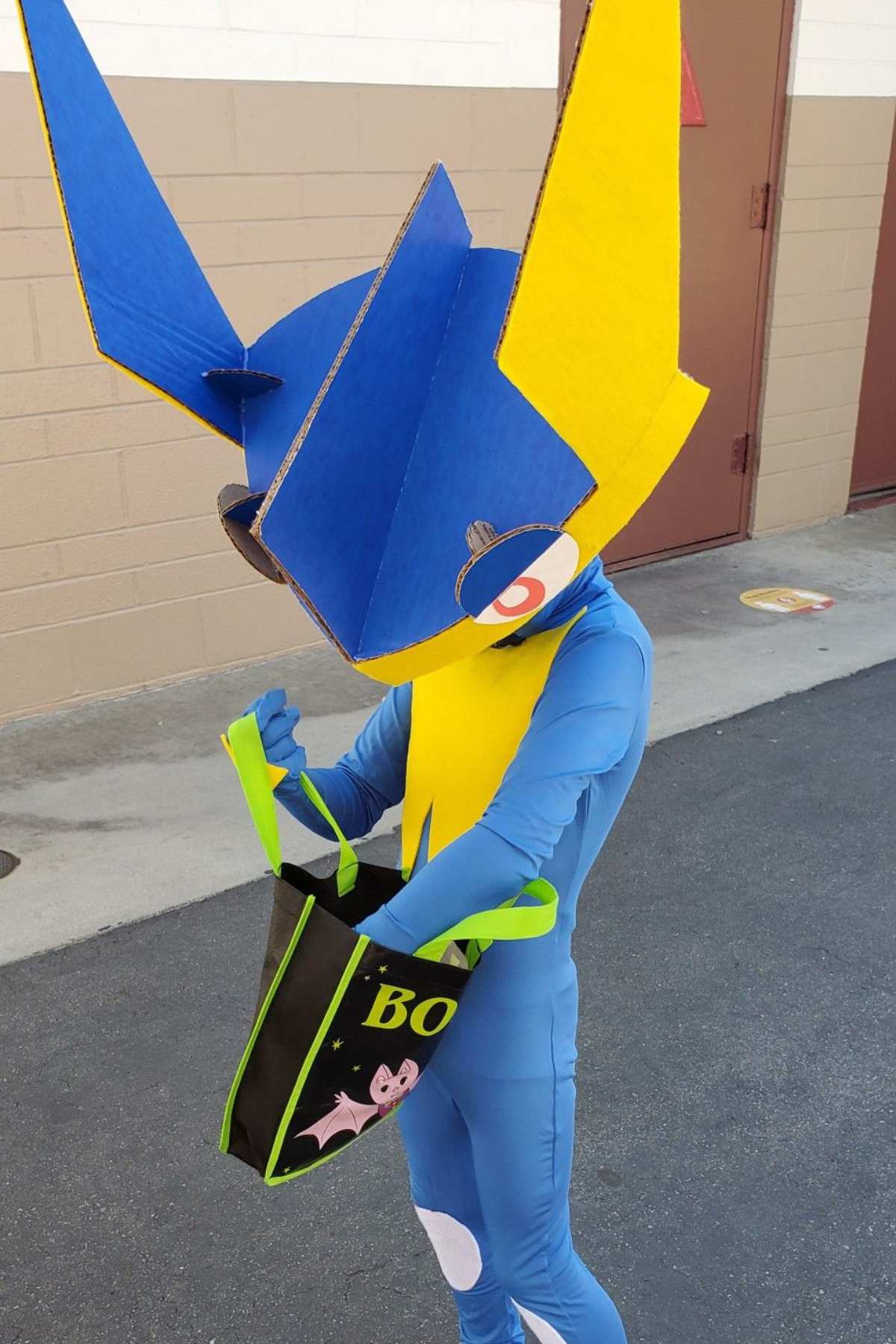 Person in a Greninja costume with a large angular headpiece holds an open Halloween trick-or-treat bag while standing outside on pavement.