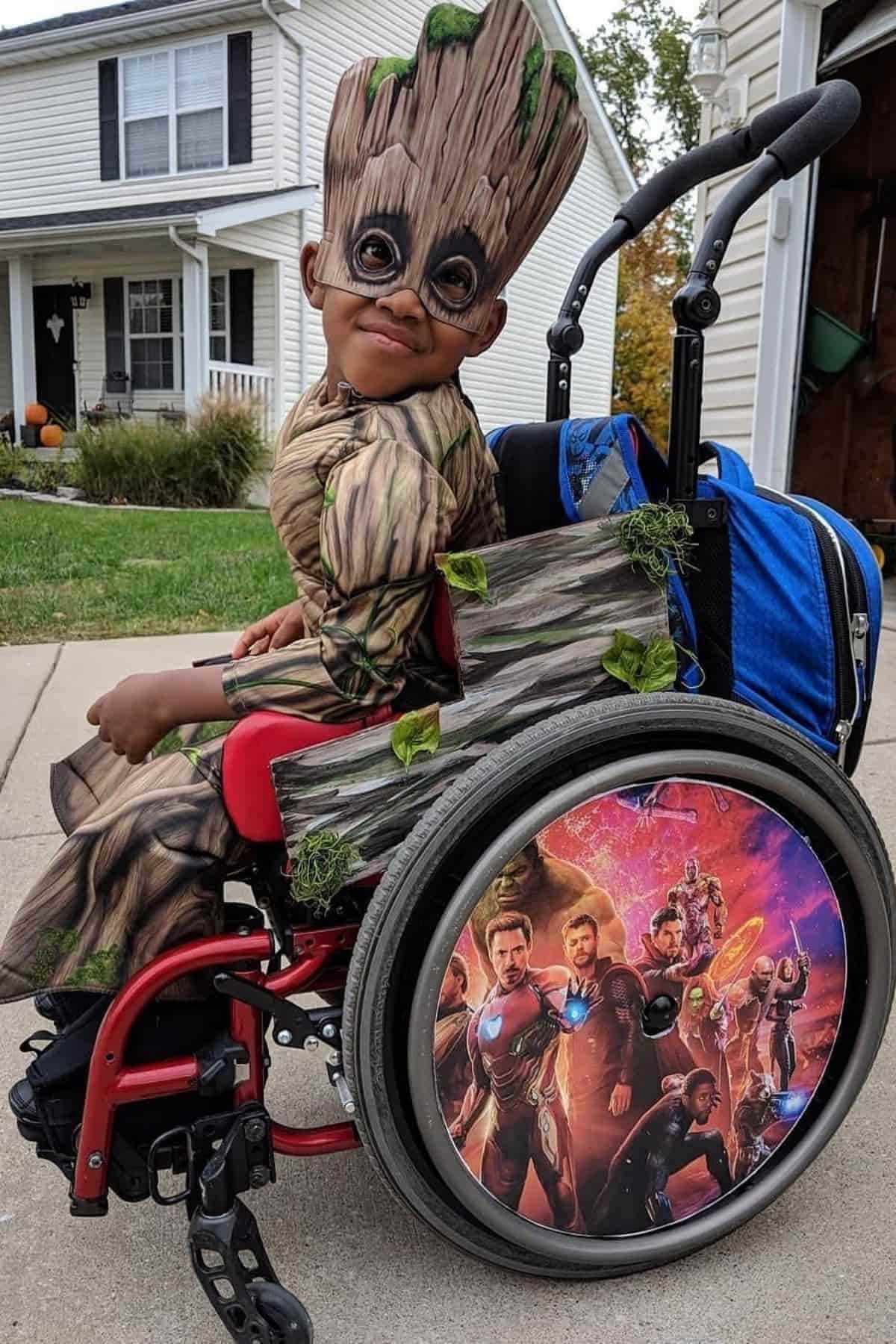 A child in a wheelchair dressed as Groot from Guardians of the Galaxy, with Avengers characters pictured on the wheelchair wheels, outdoors in a suburban neighborhood.