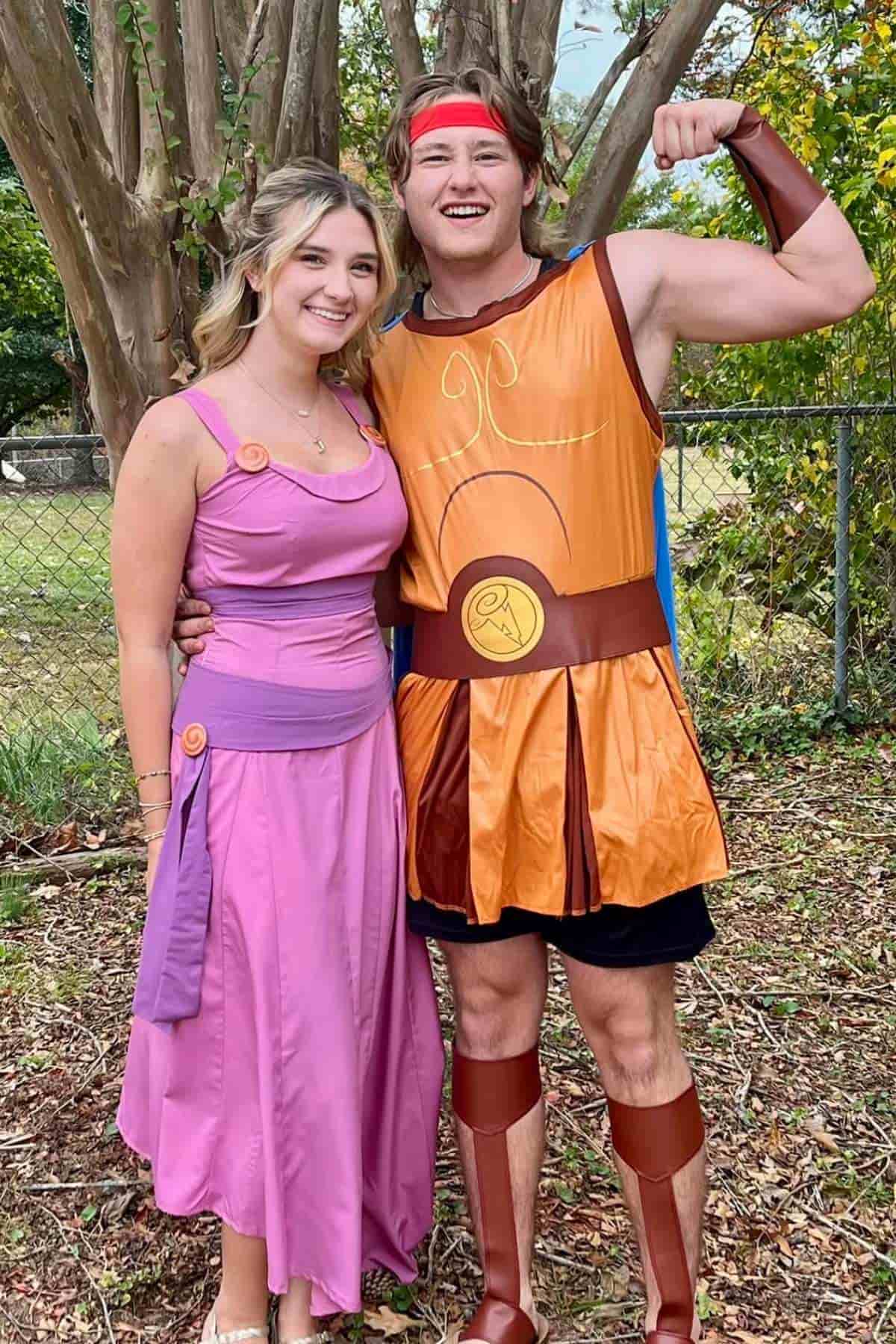 Two people pose outdoors in costumes inspired by characters from Disney’s “Hercules”; one wears a pink dress, the other a gladiator-style outfit. Trees and a chain-link fence are in the background.