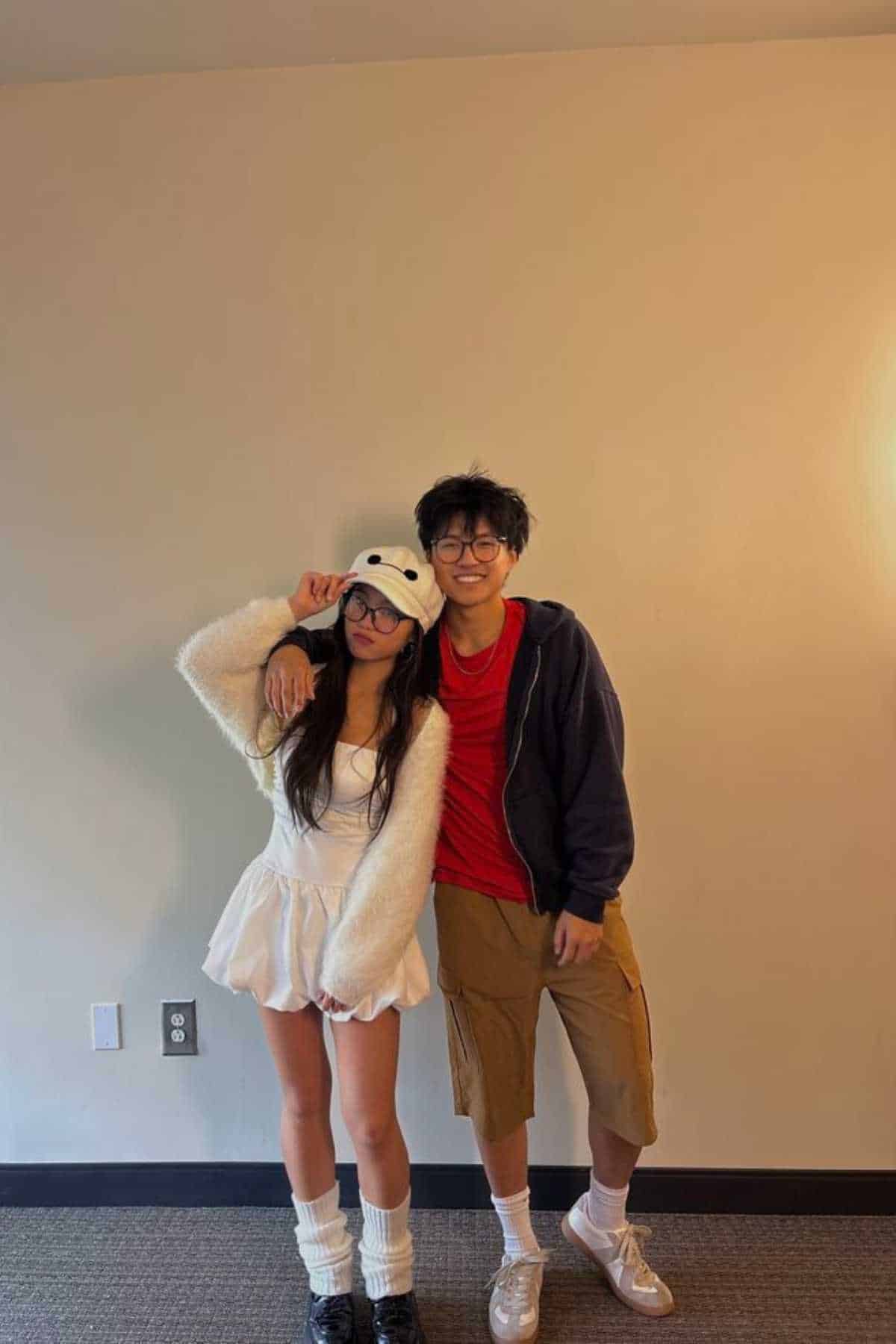 Two people stand indoors against a plain wall. The person on the left wears a white bear hat and fluffy outfit, and the person on the right wears glasses, a red shirt, a dark jacket, and tan shorts.