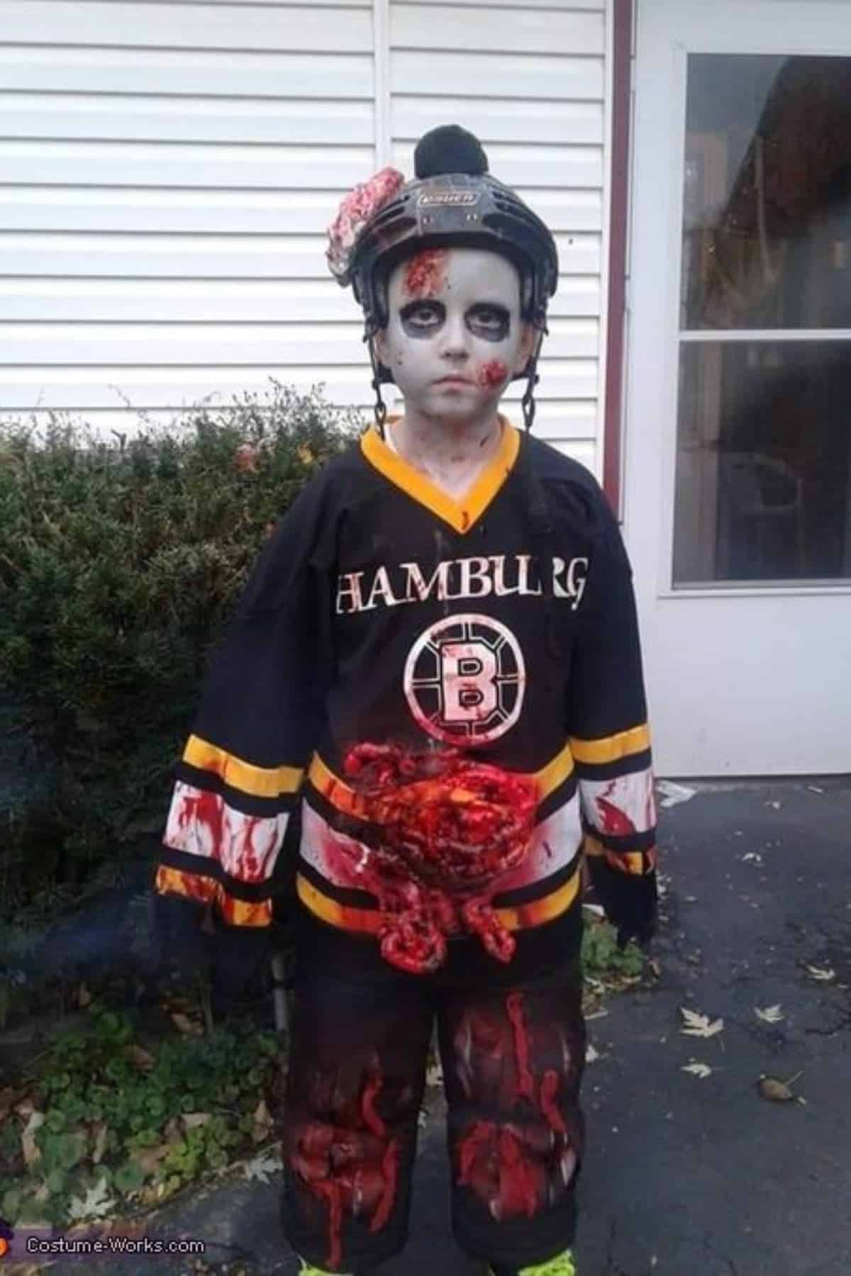 A child in a hockey uniform and helmet wears zombie makeup and fake blood, with fake intestines exposed from the shirt, standing outside near a bush and door.