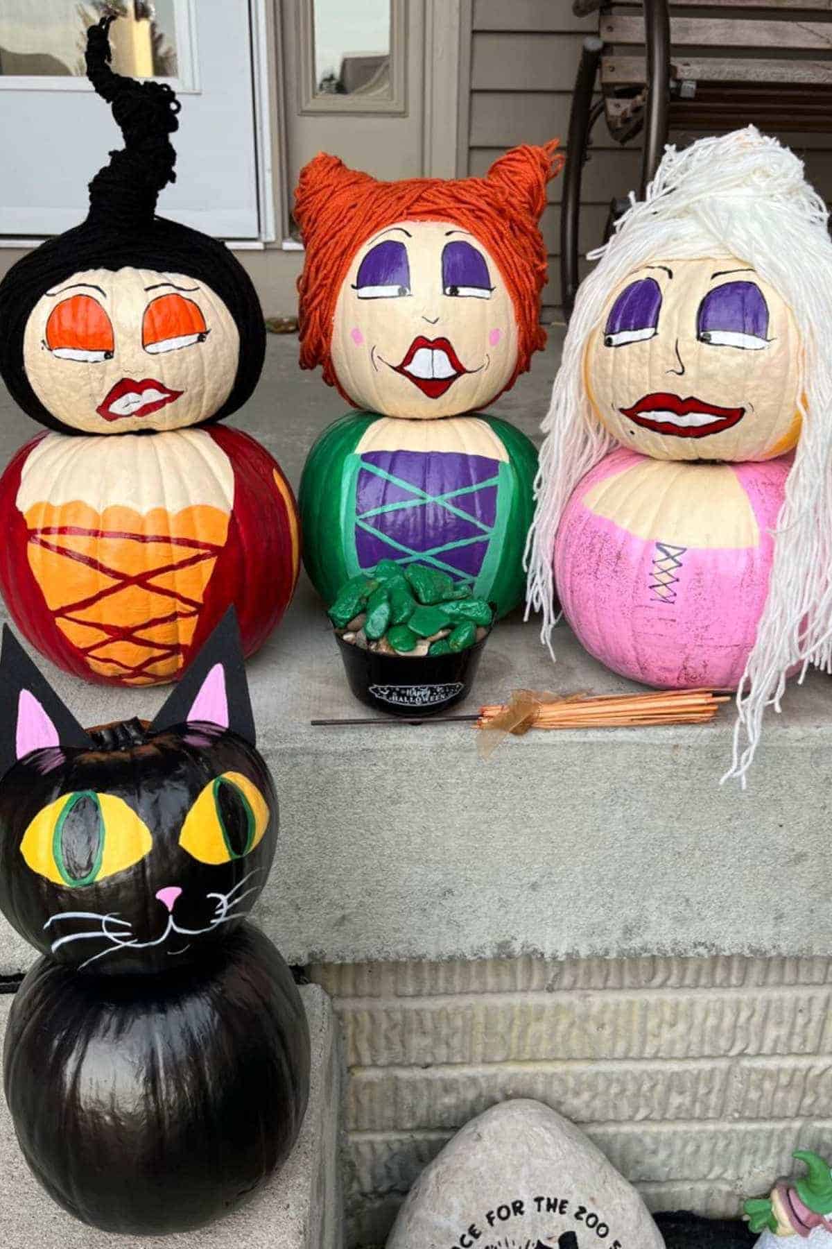 Three painted pumpkins resembling witches and a black cat pumpkin are displayed on a porch step with a small cauldron and wooden sticks.