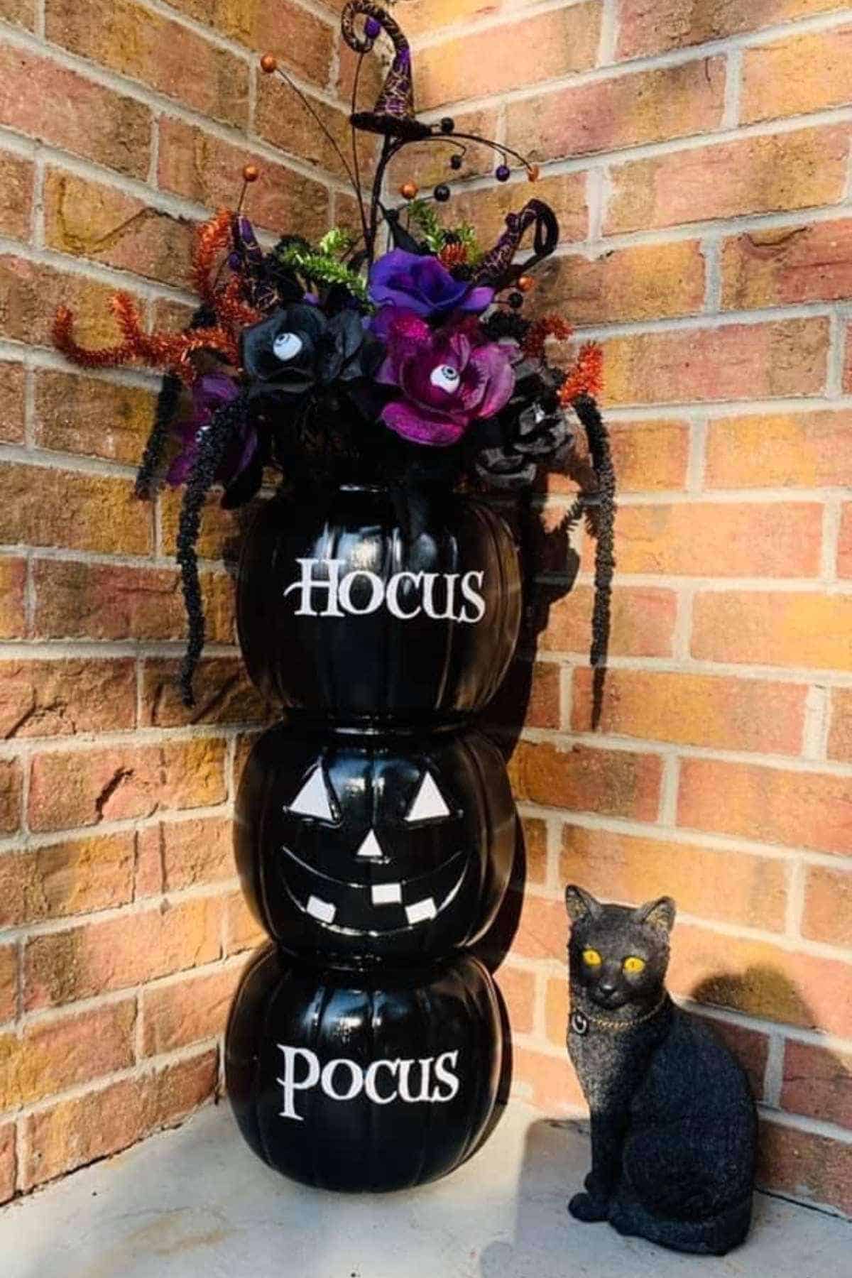 Three stacked black pumpkins with “Hocus Pocus” and a jack-o’-lantern face, topped with artificial flowers, next to a black cat statue against a brick wall.