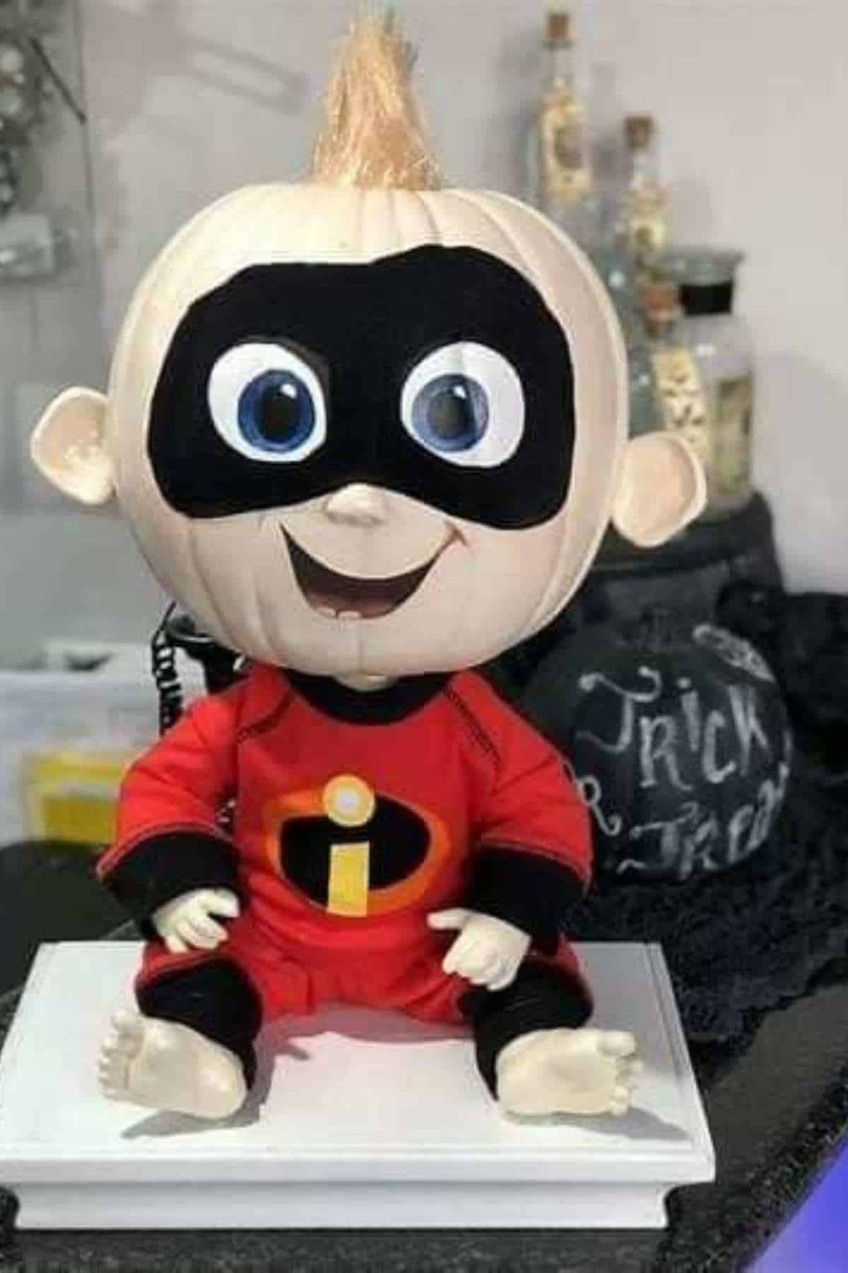 A pumpkin decorated to look like a baby in a red superhero costume with a black eye mask, resembling a character from The Incredibles, is displayed indoors.