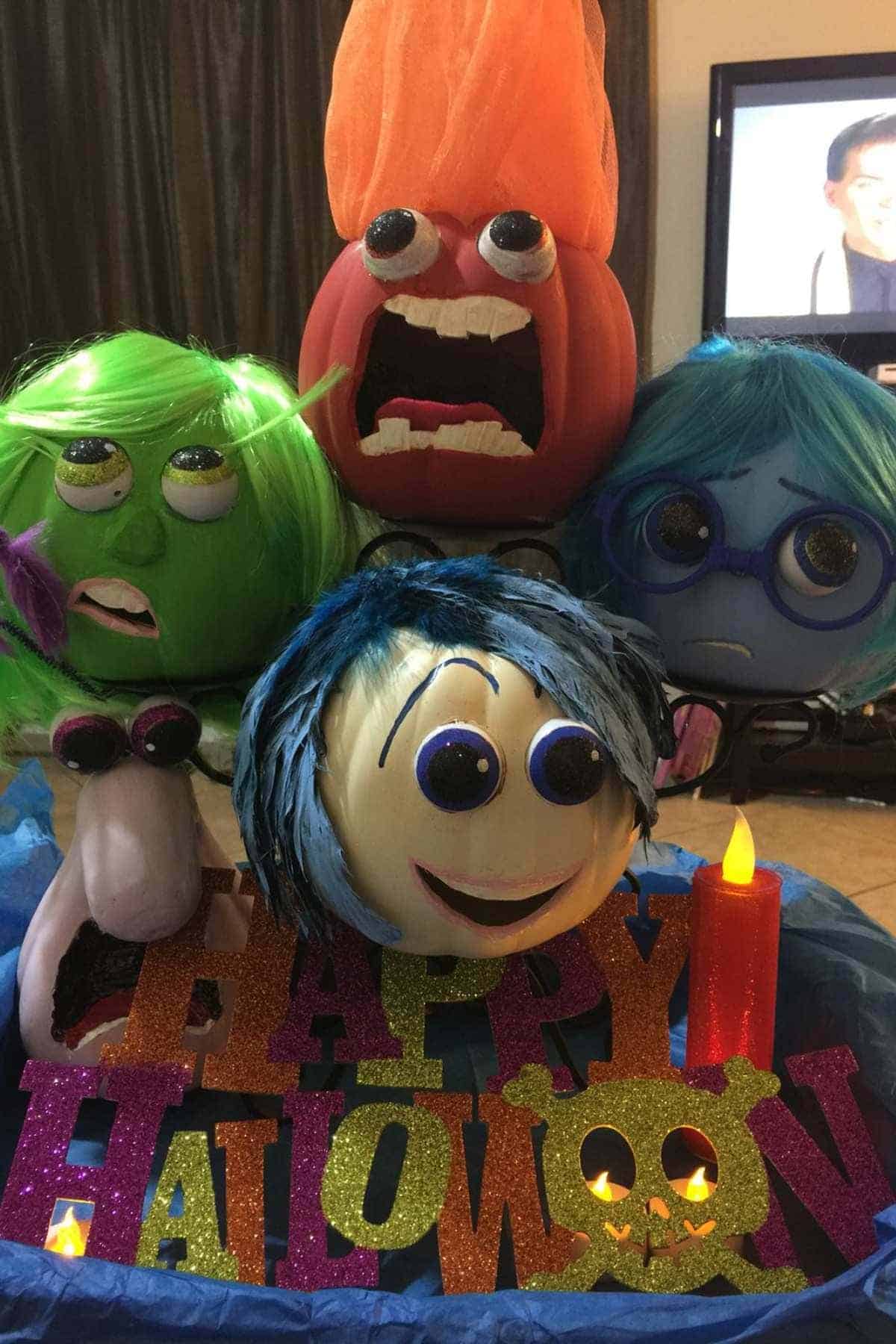 Four decorated pumpkins resembling characters from “Inside Out” are arranged behind a glittery “Happy Halloween” sign with fake candles in front.