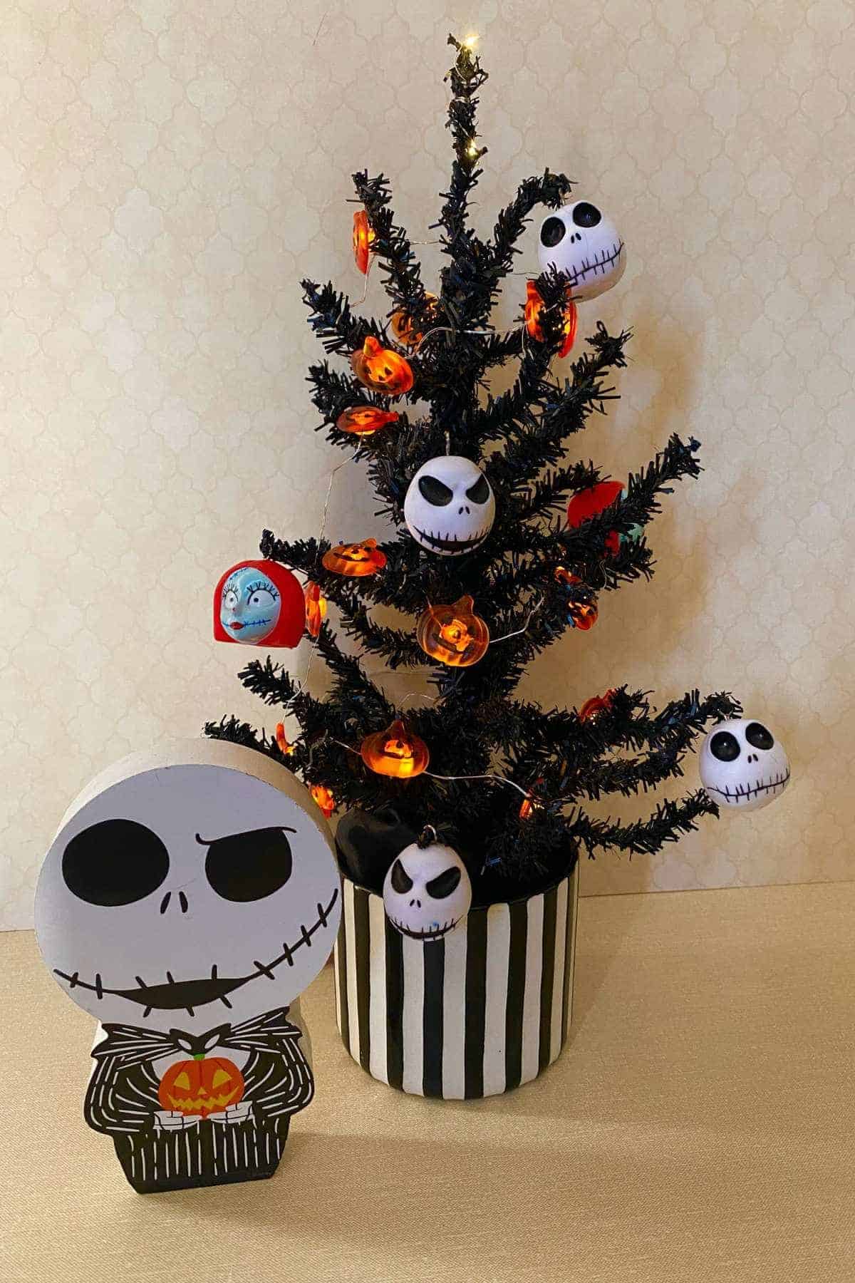 A small black Christmas tree decorated with Jack Skellington and pumpkin ornaments stands in a black and white striped pot, with a Jack Skellington figure holding a pumpkin in front.