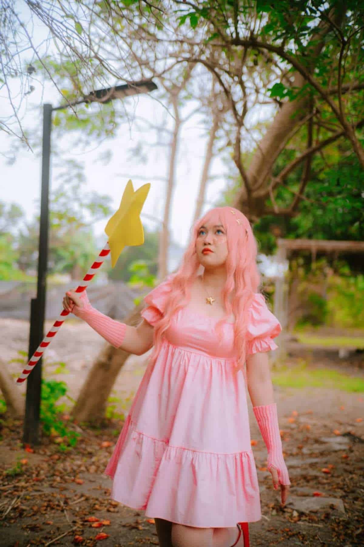 Person in a Kirby costume, wearing pink dress and pink wig holds a yellow star wand outdoors, standing among trees and greenery.
