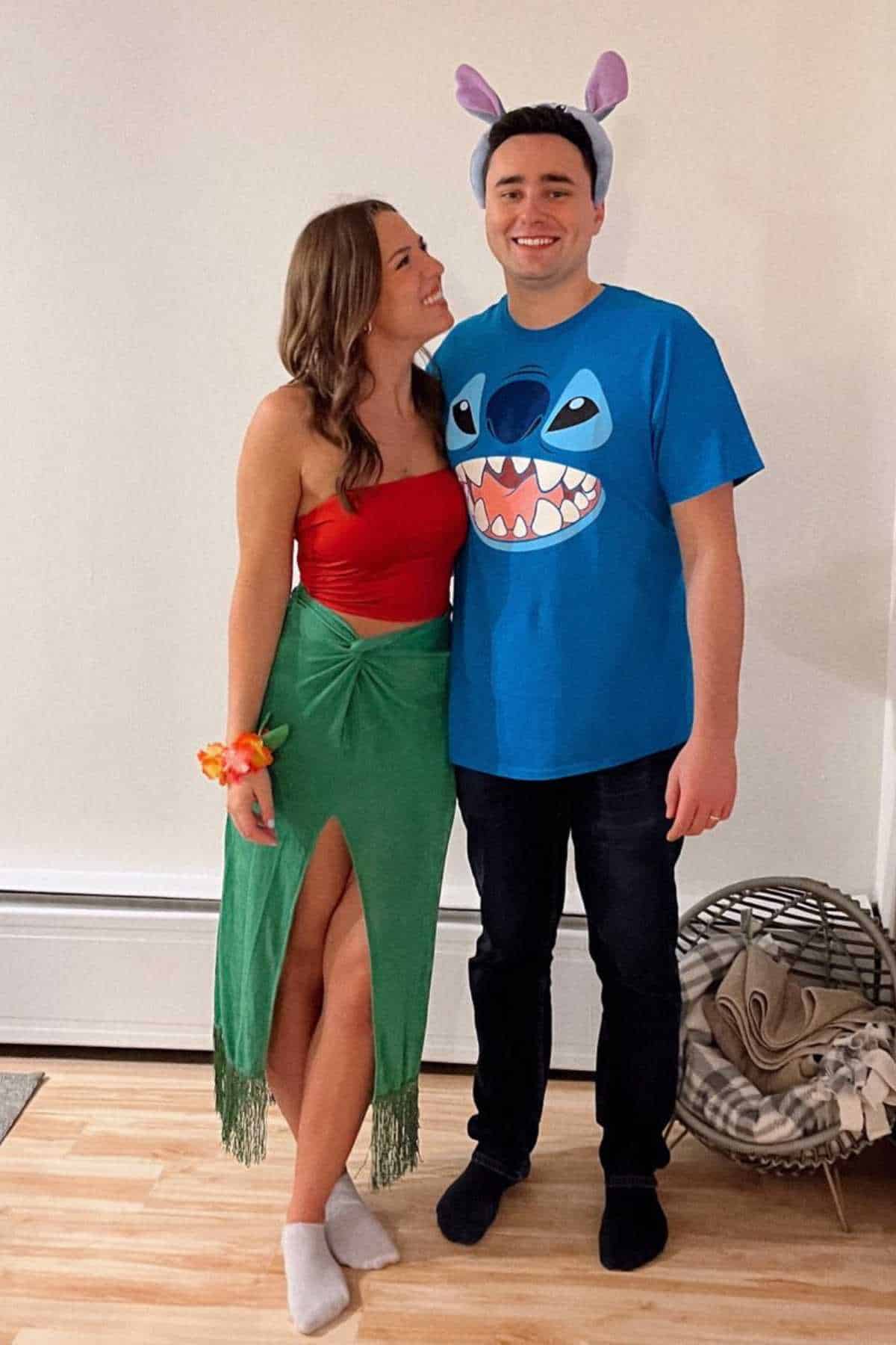 Two people in costumes stand indoors; the woman wears a red top and green skirt, while the man wears a blue Stitch shirt and purple ear headband.