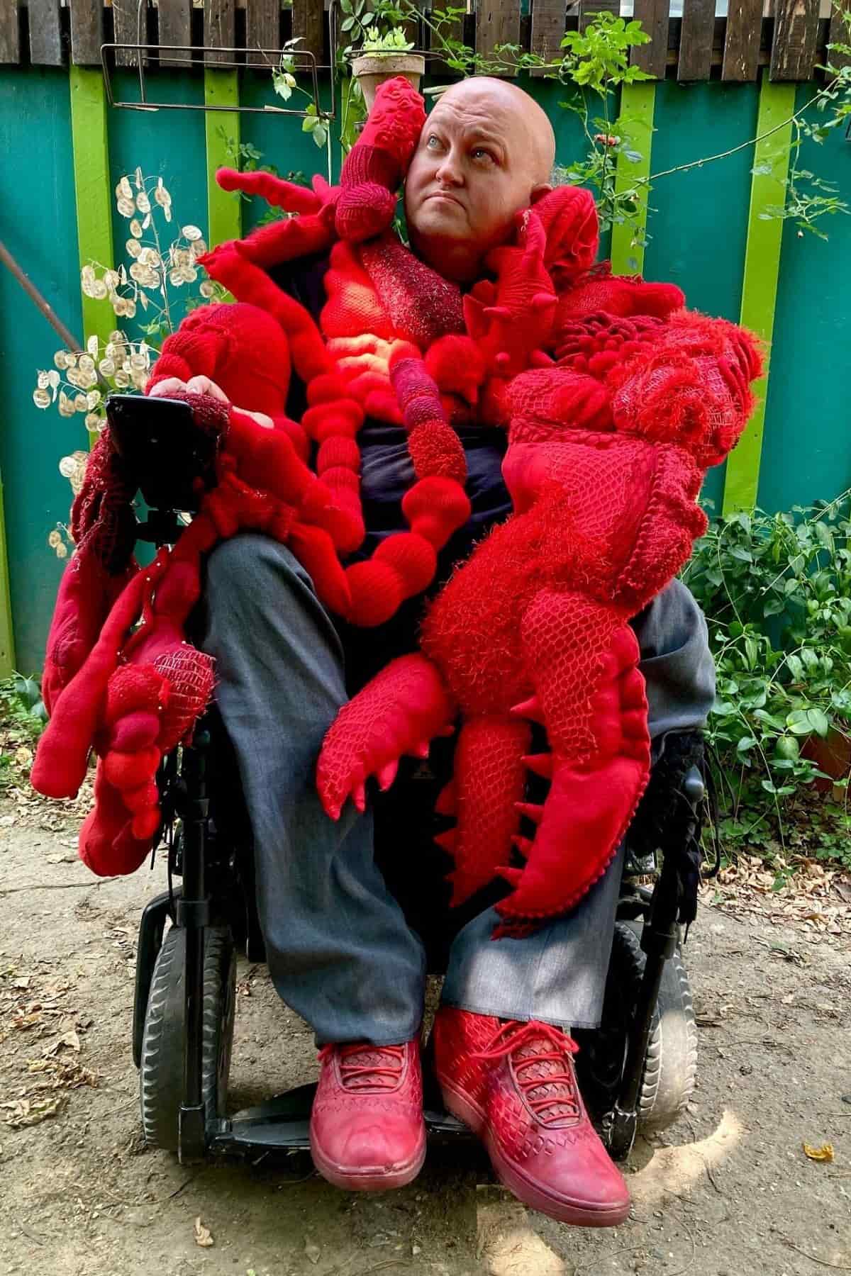 A person in a wheelchair wears a large, bright red knitted costume resembling lobsters, with a green and blue fence and plants in the background.