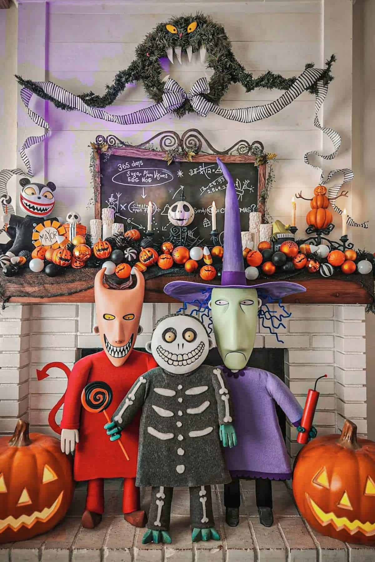 Three people in detailed costumes stand in front of a Halloween-themed fireplace decorated with pumpkins, spooky figures, and a monster garland above a chalkboard.