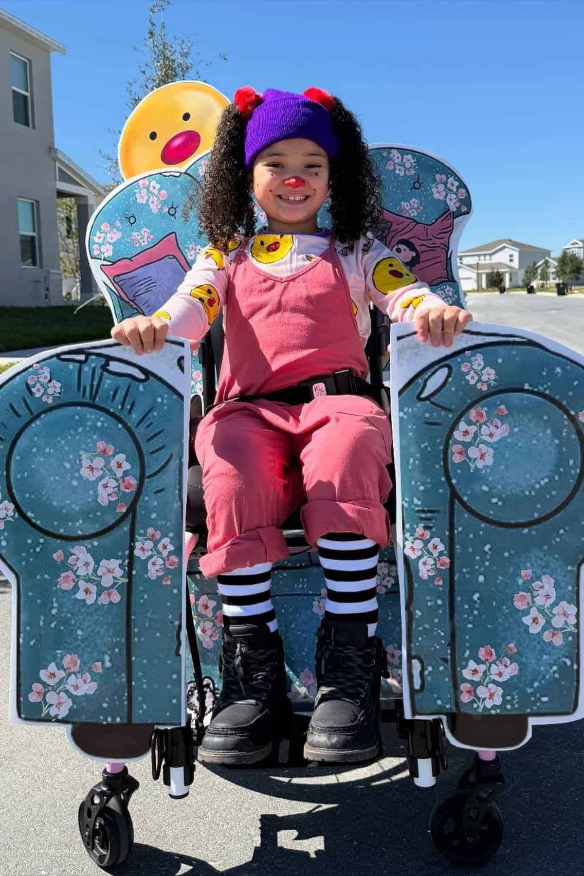 Child in a wheelchair costume decorated as a blue armchair with floral patterns, dressed in colorful clothes and a purple hat, smiling outdoors on a sunny day.