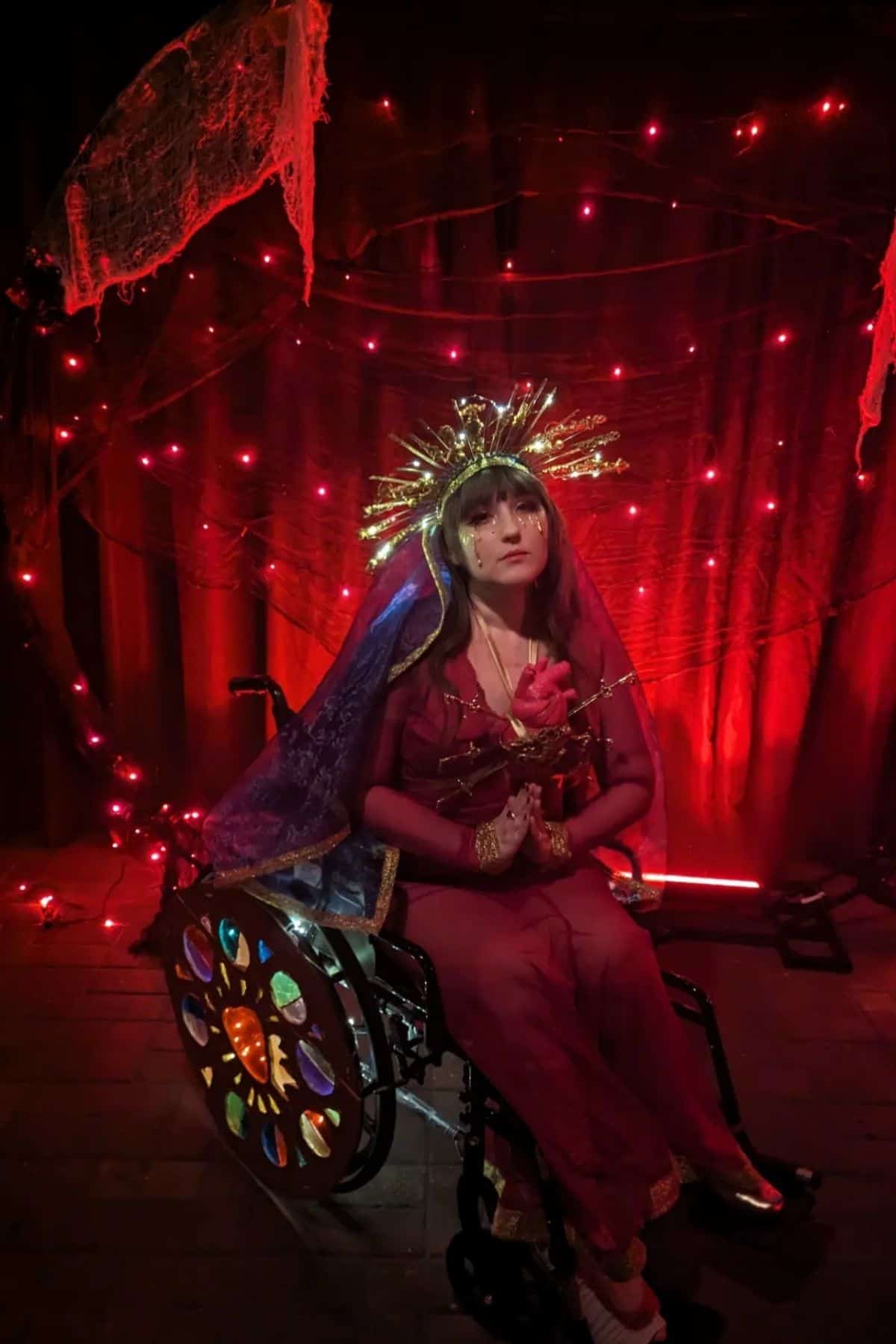 A person in a wheelchair wears a gold headpiece, red outfit, and blue veil, holding a rose, posed against a backdrop of red lights and draped fabric.
