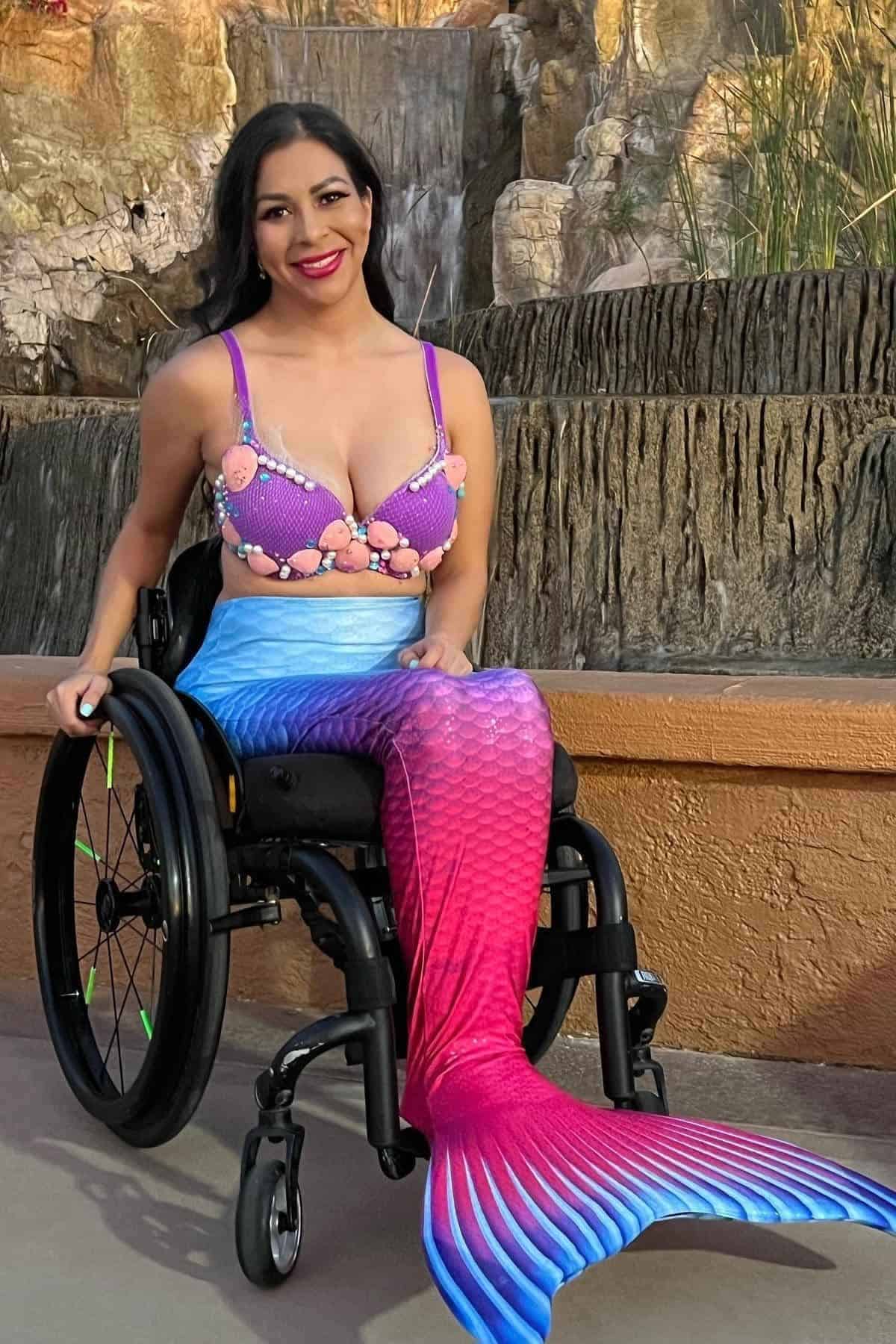 A woman in a wheelchair wears a colorful mermaid costume with a blue and pink tail and a seashell top, sitting outdoors in front of a waterfall.