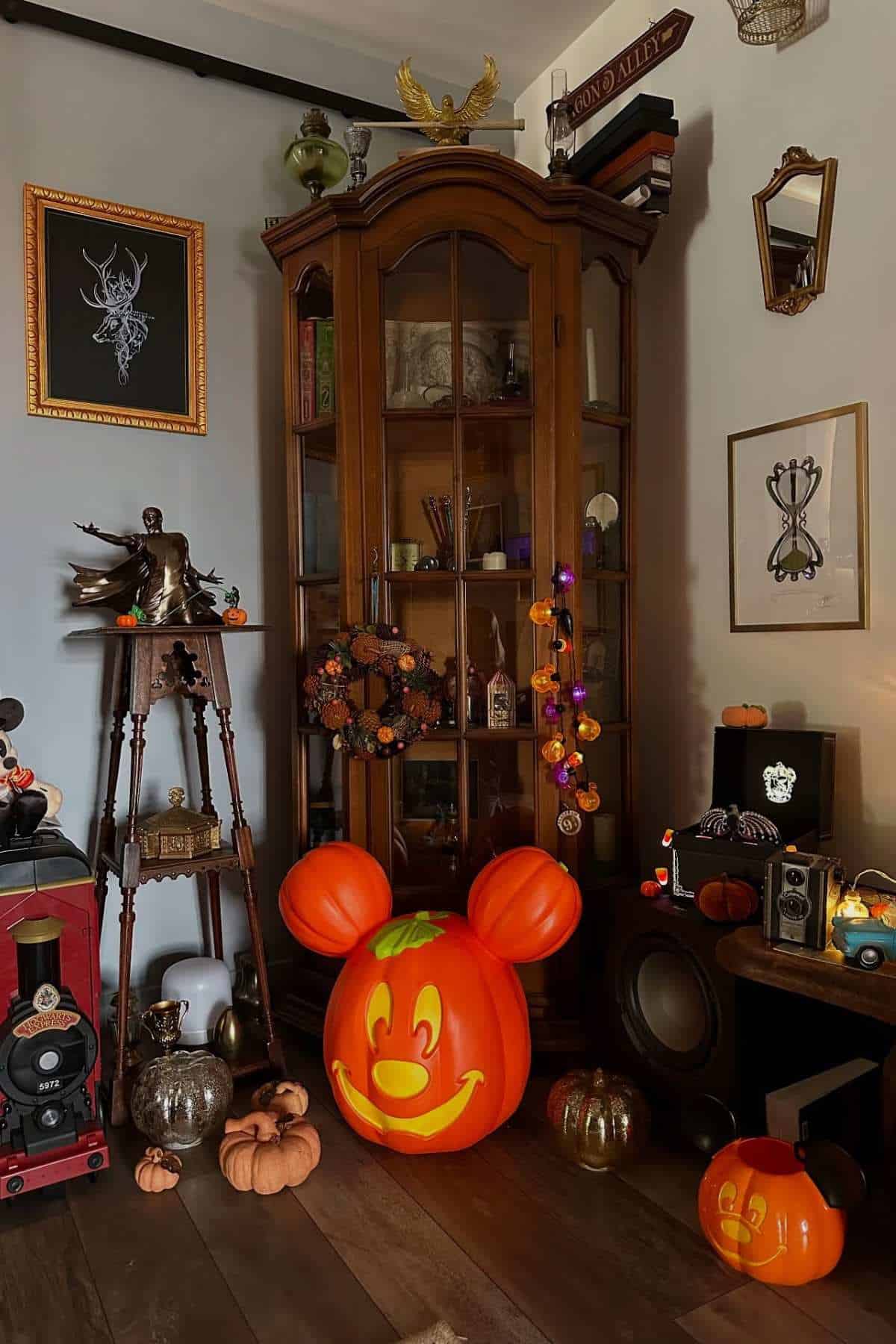 Room corner decorated for Halloween with pumpkins, wreaths, lights, and vintage items. A large Mickey Mouse pumpkin sits on the floor next to a wooden cabinet filled with collectibles.
