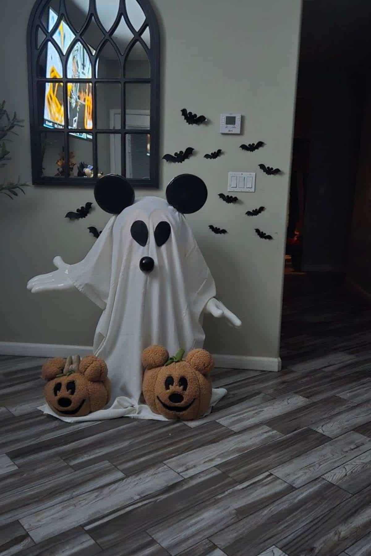 A ghost costume with Mickey Mouse ears stands by the wall, flanked by two pumpkin decorations with Mickey faces; black bat decals are on the wall behind.