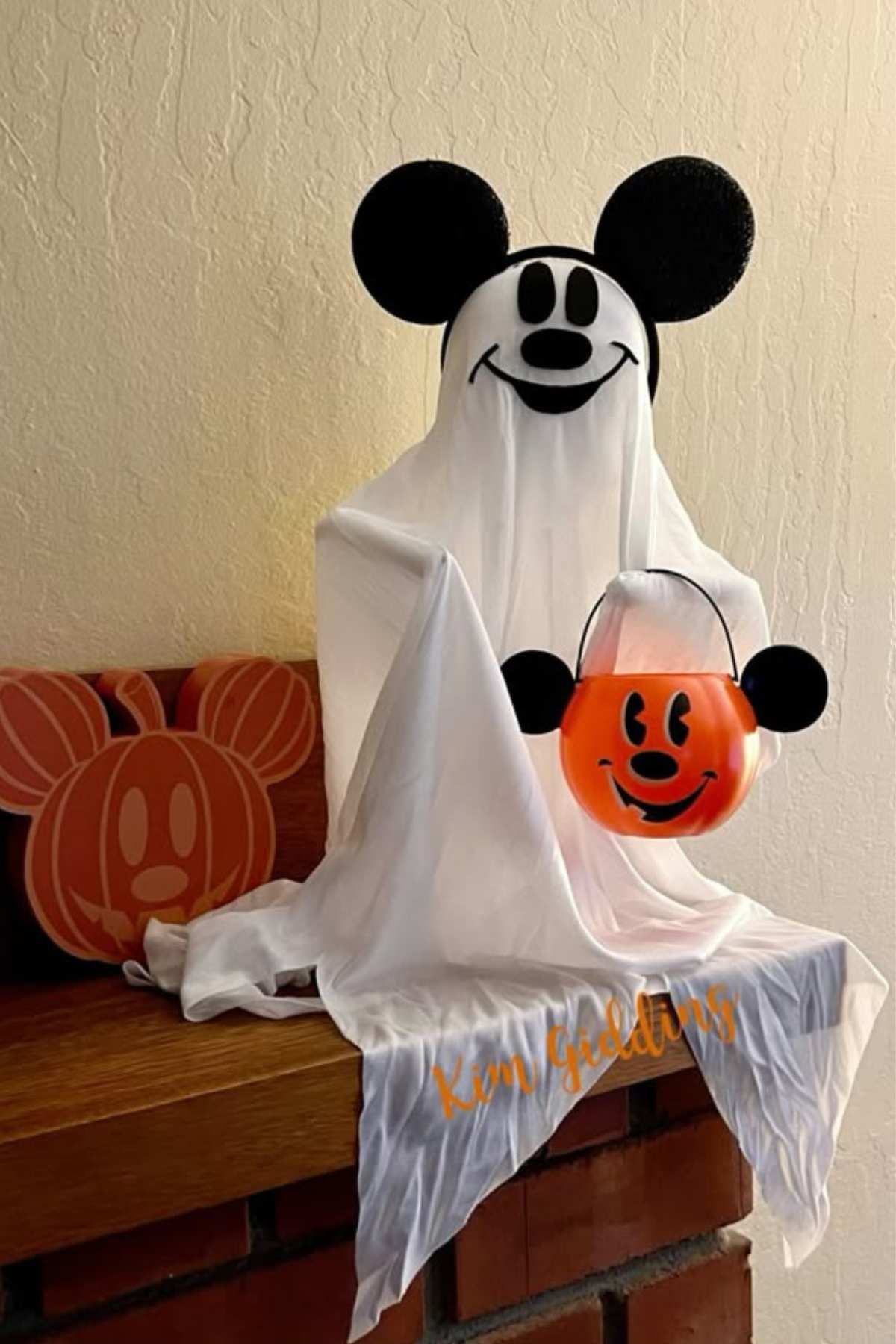 A ghost costume with Mickey Mouse ears sits on a fireplace next to a pumpkin decoration, holding an orange Mickey Mouse pumpkin bucket.