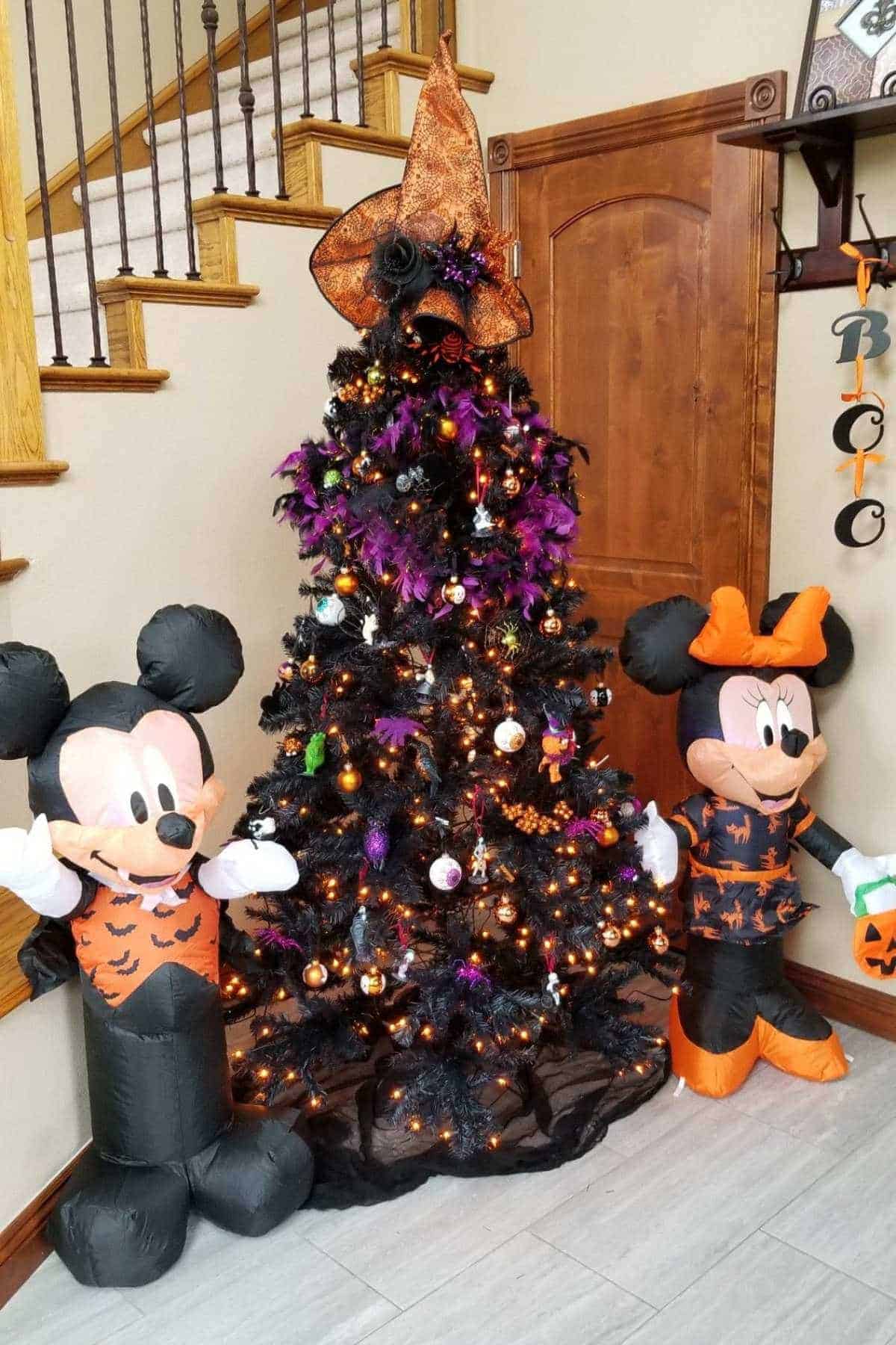 A black Halloween tree with orange and purple lights stands between inflatable Mickey and Minnie Mouse decorations in Halloween costumes.