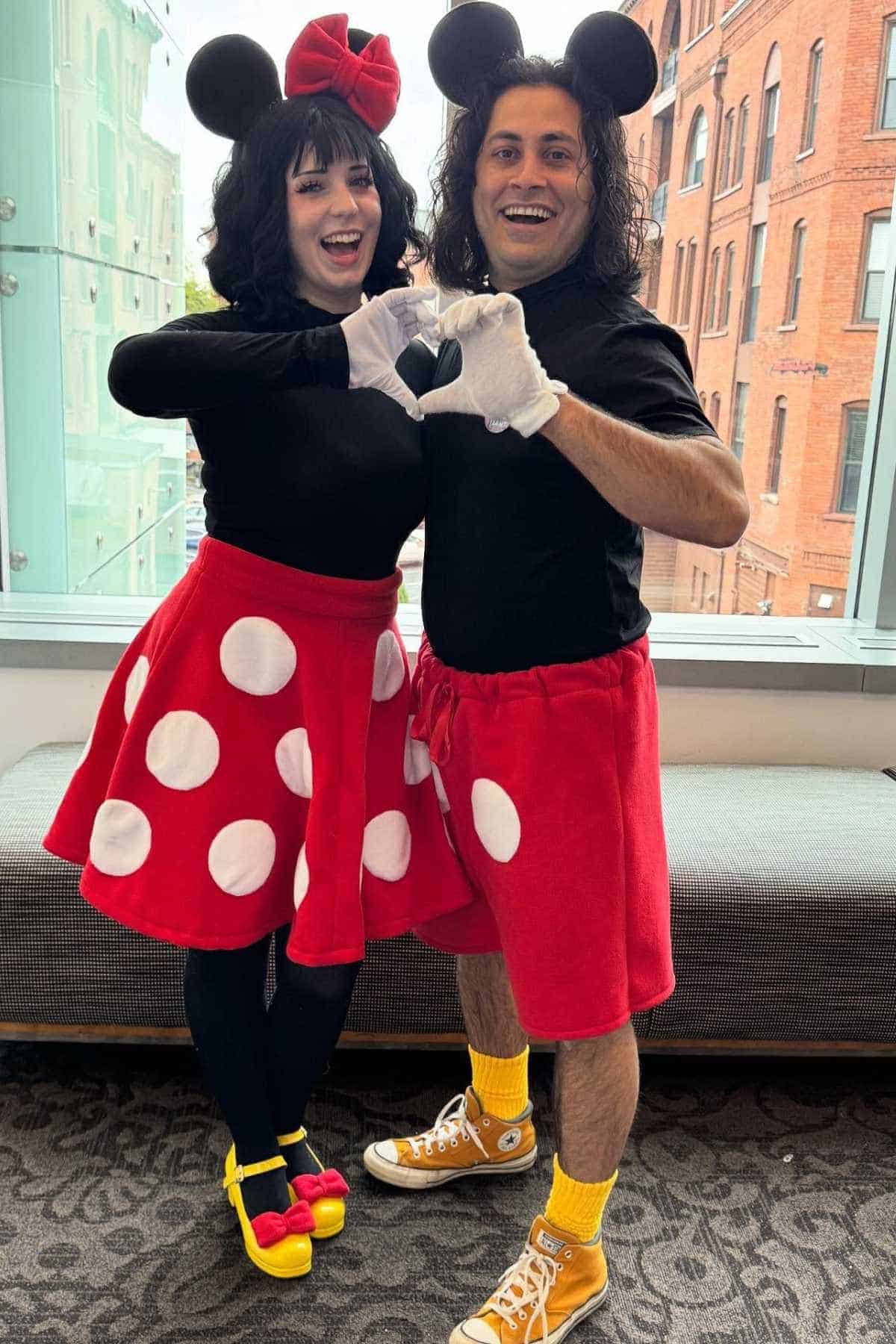 Two people dressed as Minnie and Mickey Mouse stand indoors, smiling and forming a heart shape with their gloved hands.