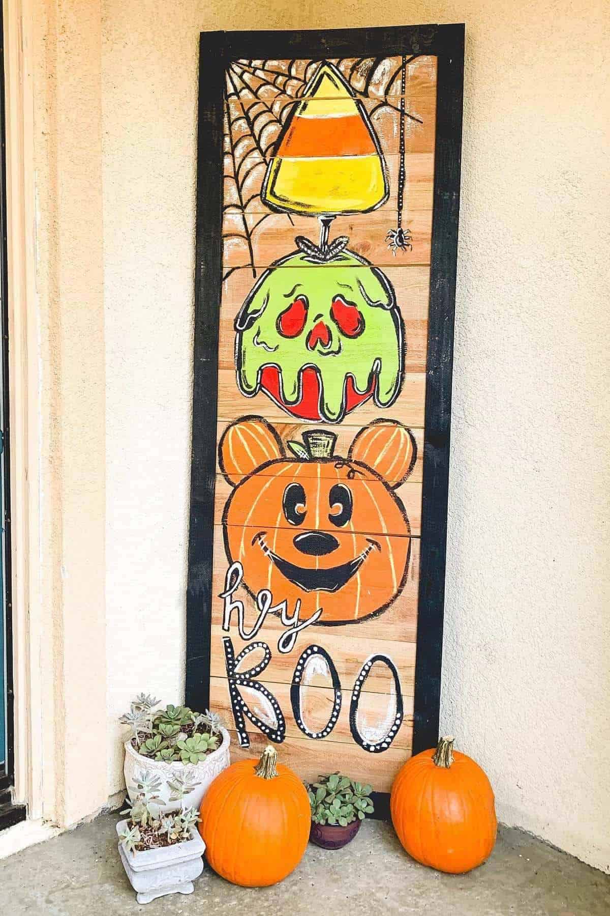A Halloween-themed wooden sign with a candy corn, green skull, and orange bear face, with “hi BOO” text. Three pumpkins and potted succulents sit at the base.