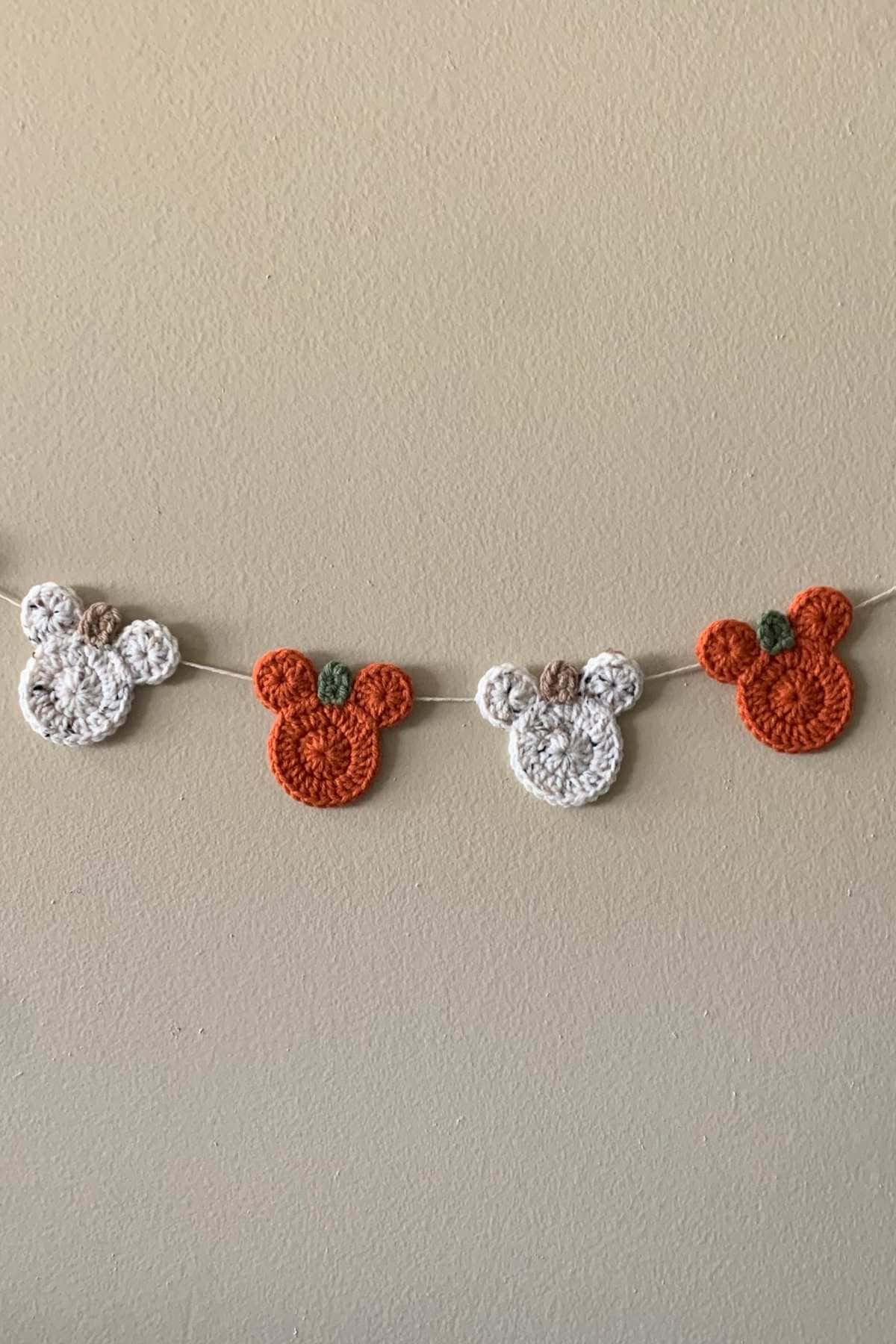 Four crocheted Mickey Mouse-shaped ornaments, two white and two orange, hang on a string against a beige wall. The orange ones have small green details resembling pumpkin stems.