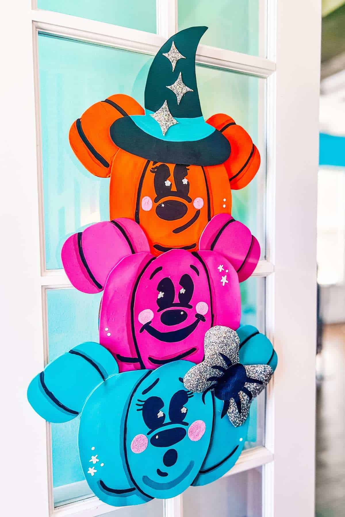 Three stacked Mickey Mouse-shaped pumpkins in orange, pink, and blue, decorated with painted faces, a witch hat, and a glittery bow, hang on a glass door.
