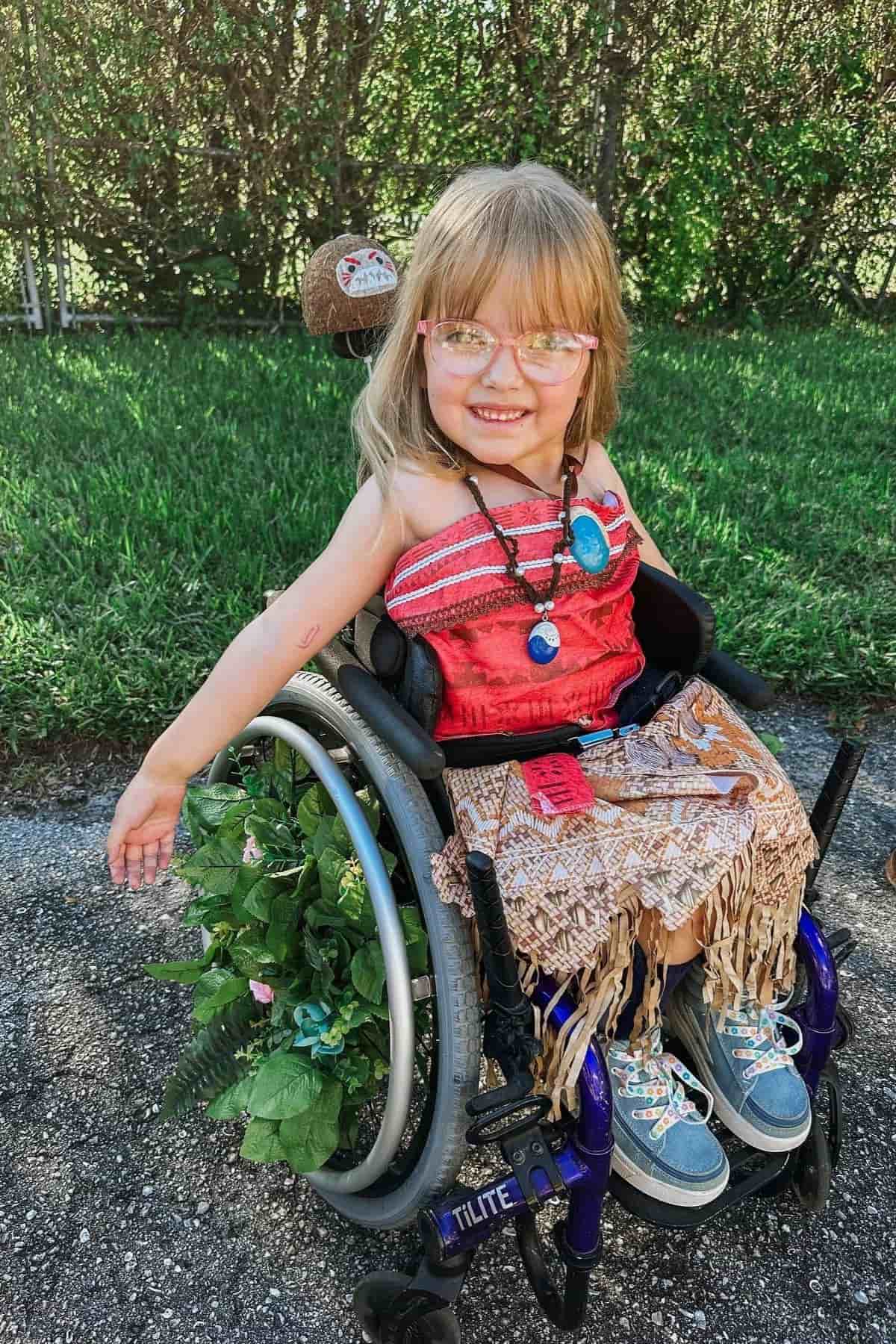 Young girl in a wheelchair dressed in a red and tan costume with a grass skirt, necklaces, and glasses, smiling outdoors on a sunny day.