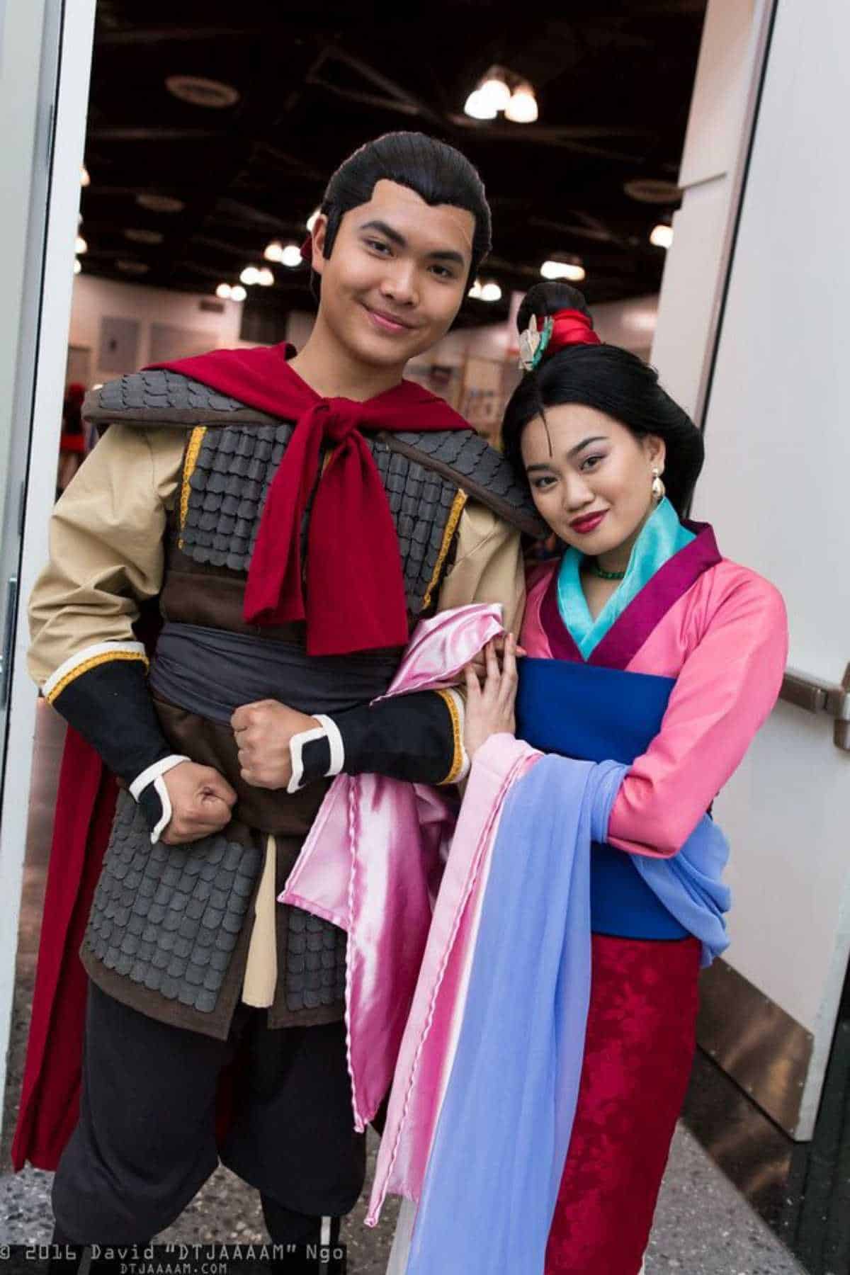 Two people in detailed cosplay costumes pose together indoors, one dressed as a warrior with armor and a red cape, the other as a woman in a pink and blue traditional-style dress.