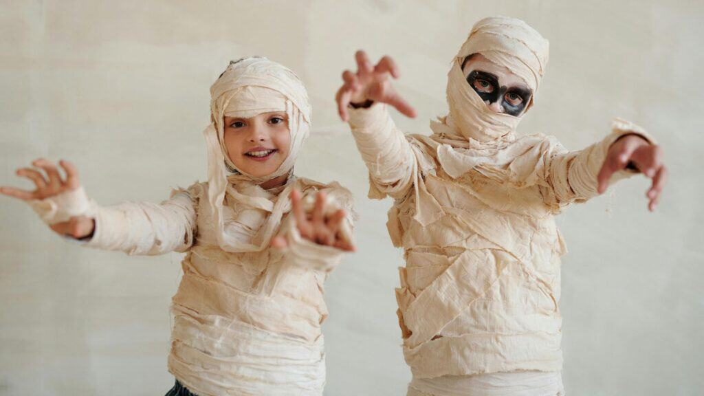 Two children dressed as mummies in beige wrappings stand with arms raised and reaching forward. One has dark face paint around the eyes. Neutral background.