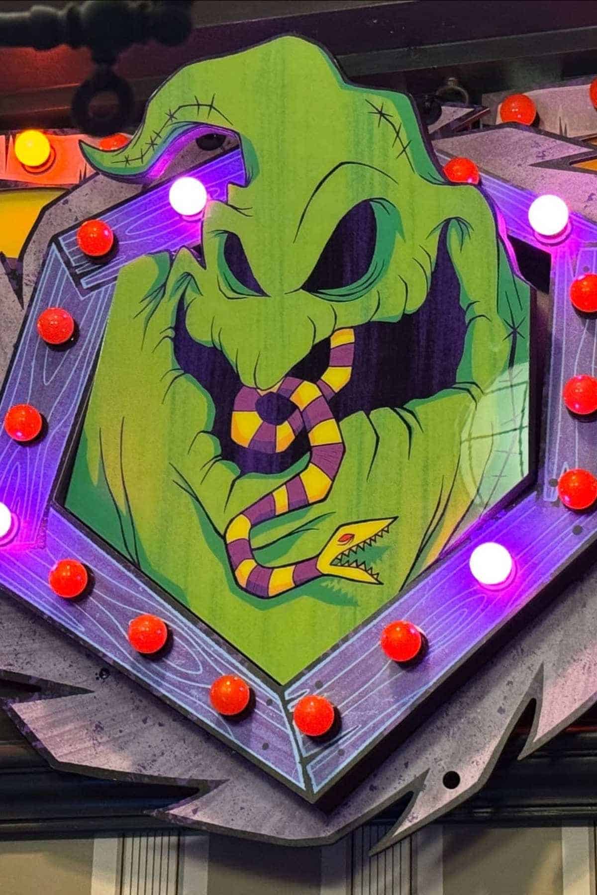 Illustration of a green, ghostly figure with hollow eyes and mouth, holding a striped snake in its mouth, surrounded by red and pink light bulbs.