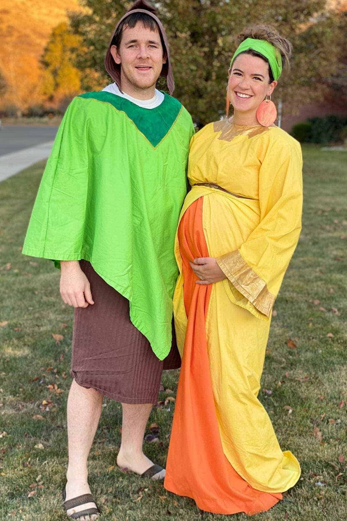 Two adults stand on grass wearing costumes inspired by characters from “The Emperor’s New Groove.” The woman is visibly pregnant and both are smiling at the camera.