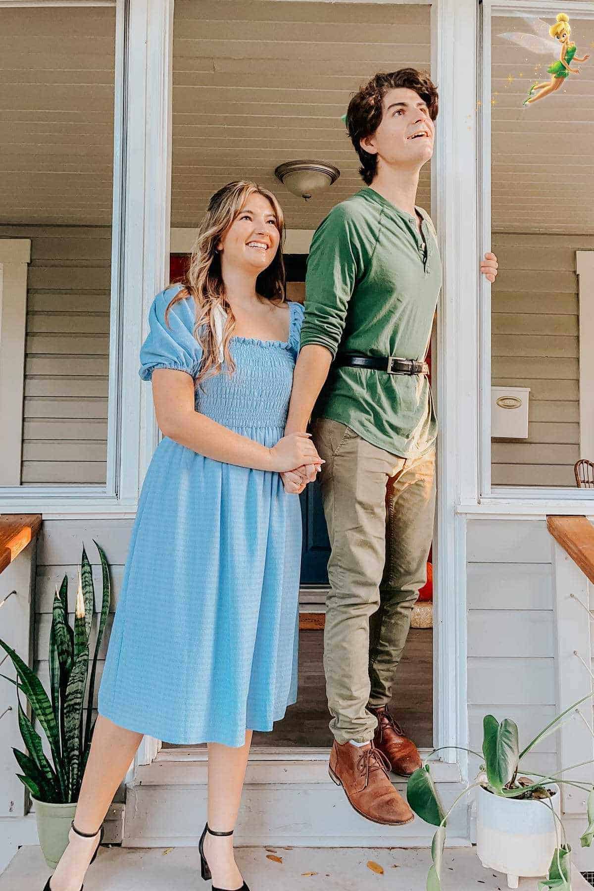 Two people dressed as Wendy and Peter Pan stand on a porch holding hands, looking up and smiling; a small image of Tinker Bell is flying above them.