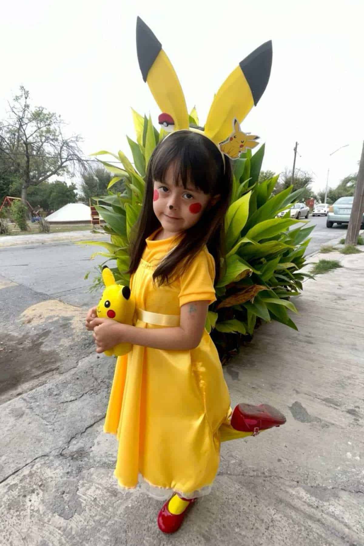 Young girl in a yellow Pikachu costume with red shoes poses on a sidewalk, holding a Pikachu plush toy, with green bushes and a street in the background.