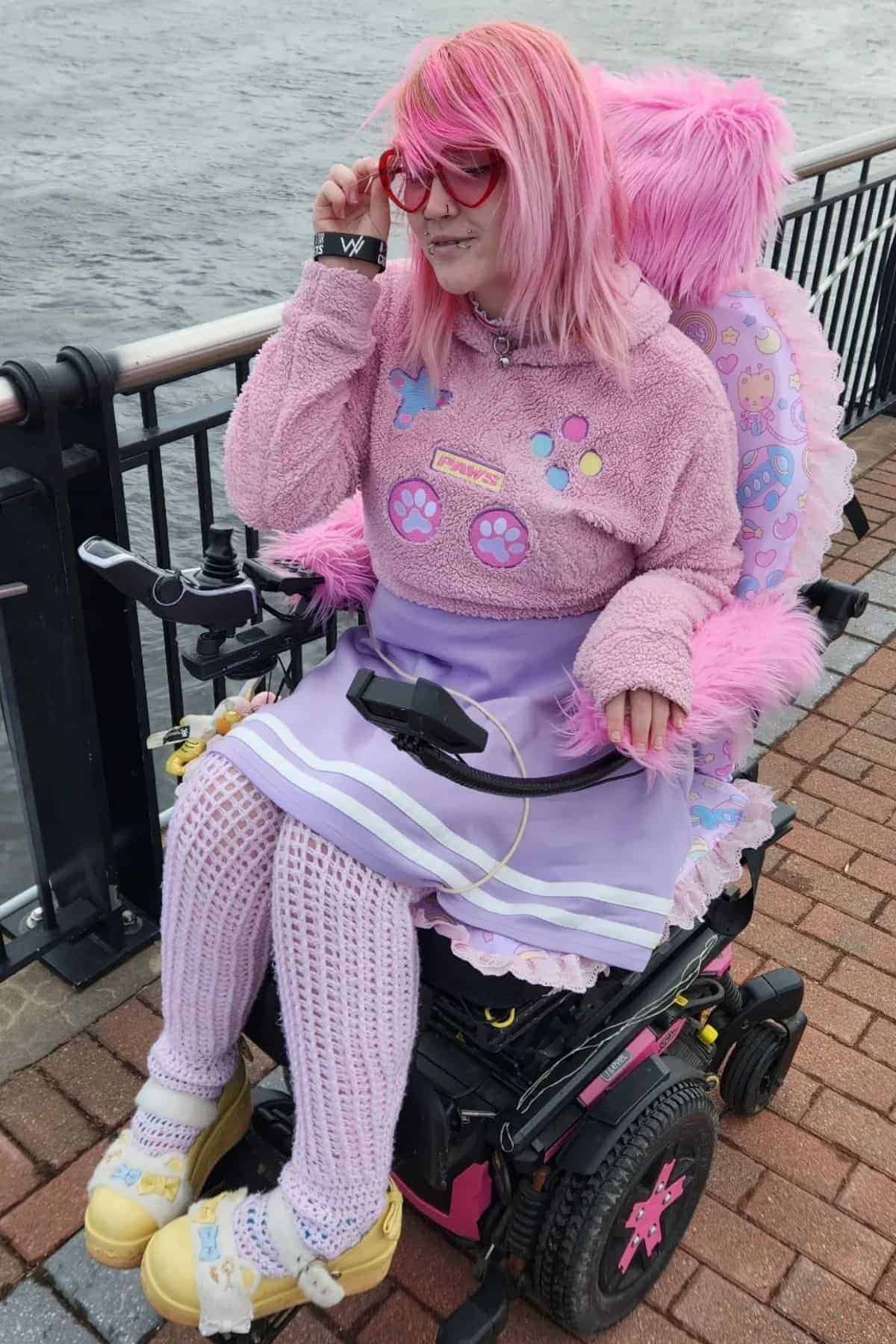 A person with pink hair and pink sunglasses sits in a decorated motorized wheelchair by a waterfront, wearing a pink and purple outfit with paw prints and lace, and pastel yellow shoes.