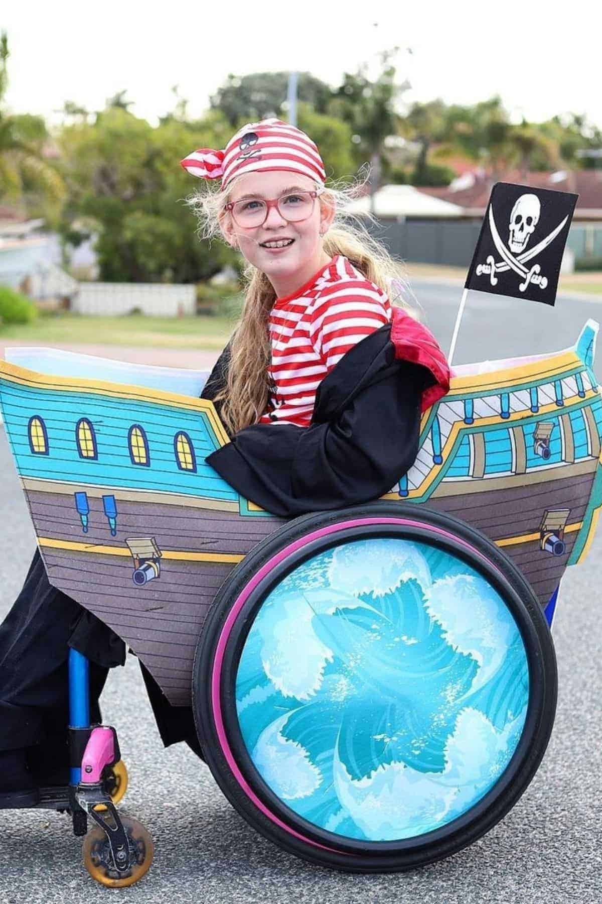 A girl in a wheelchair decorated as a pirate ship, wearing a red striped shirt, pirate hat, and glasses, smiles outdoors.