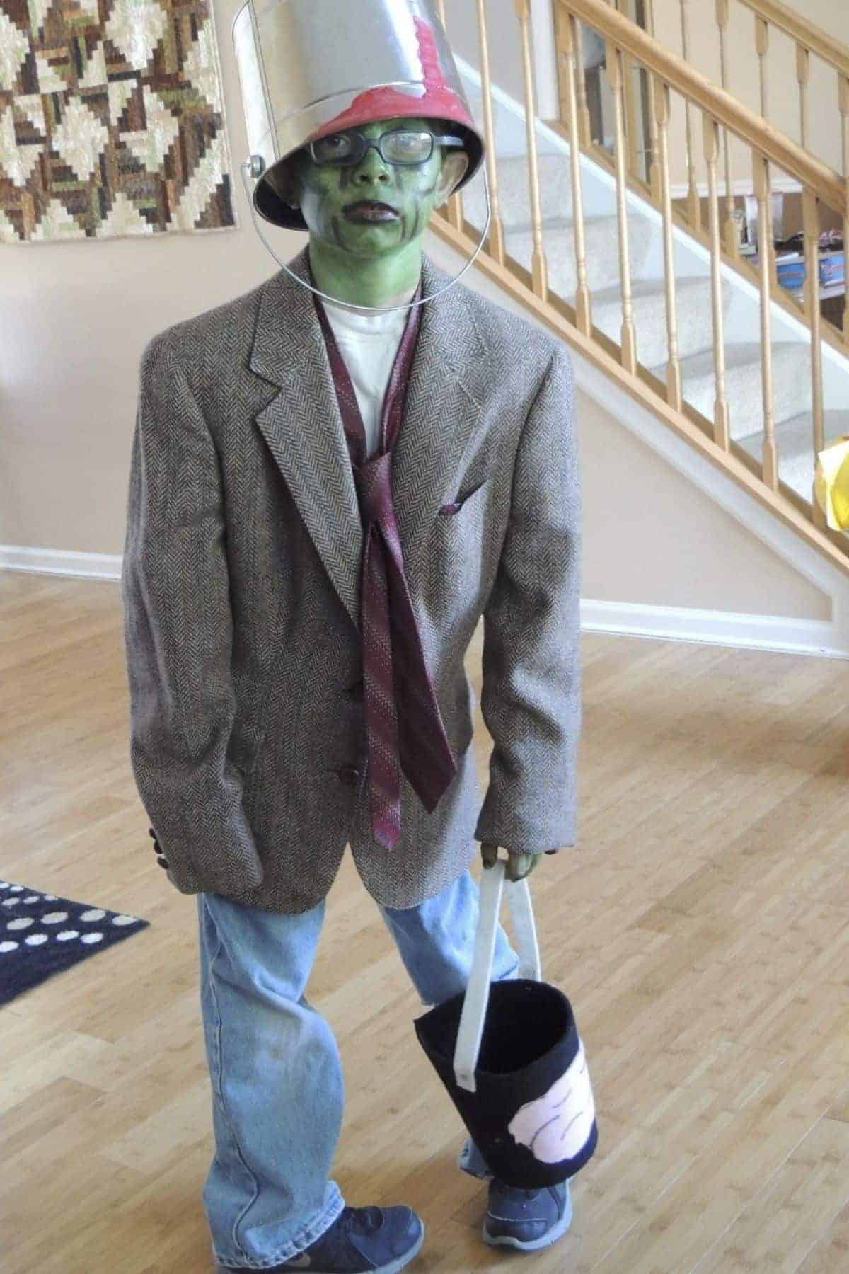 Child in oversized suit, green face paint, and bucket on head stands indoors holding a Halloween bucket.
