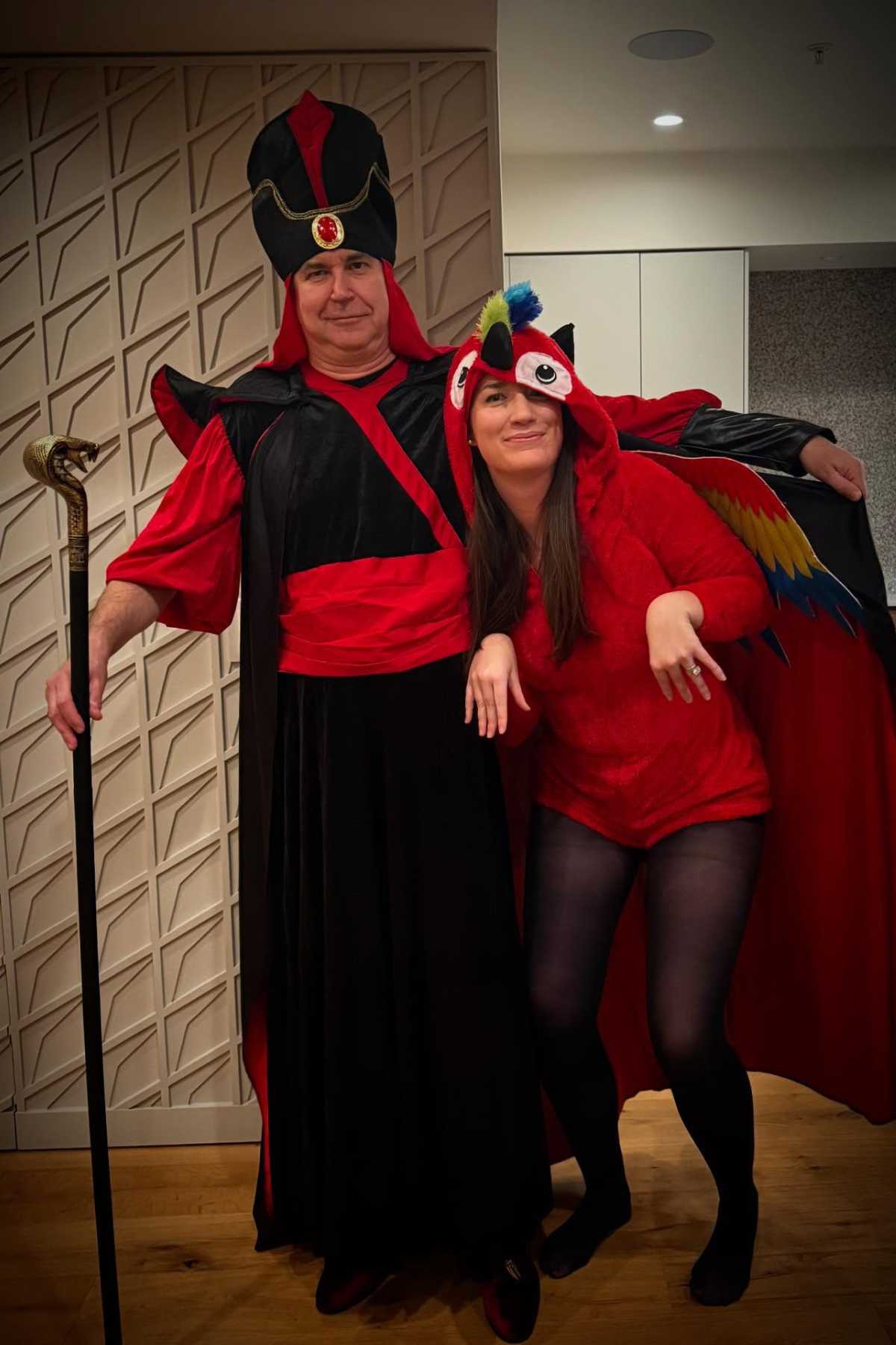 Two adults in costumes pose indoors; one is dressed as a regal character with a staff and red robe, the other as a red bird with wings and a beak hat.