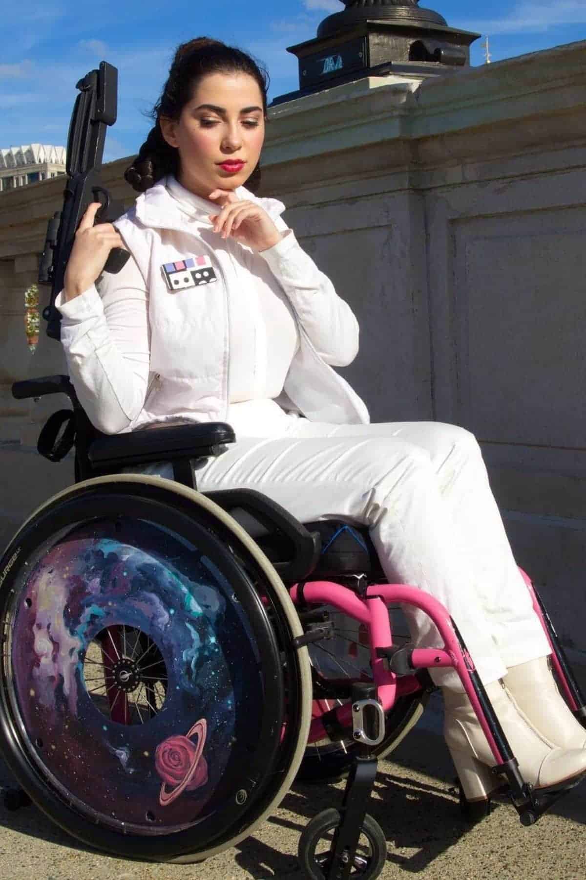 Woman in a white outfit sits in a wheelchair with galaxy-themed wheel covers, holding a prop blaster, posing outdoors by a stone railing.