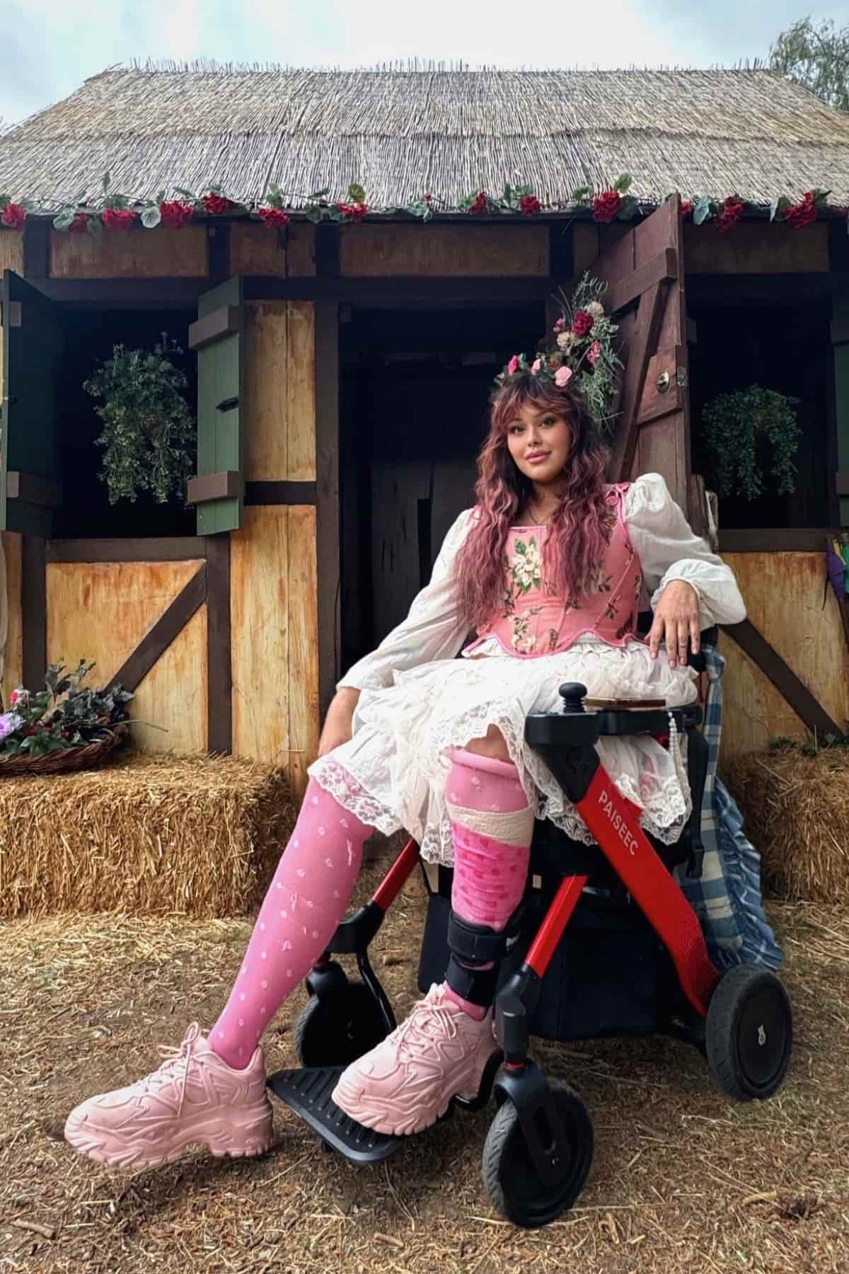 A woman in a pink dress and floral crown sits in a red wheelchair in front of a rustic building with hanging plants and a thatched roof.