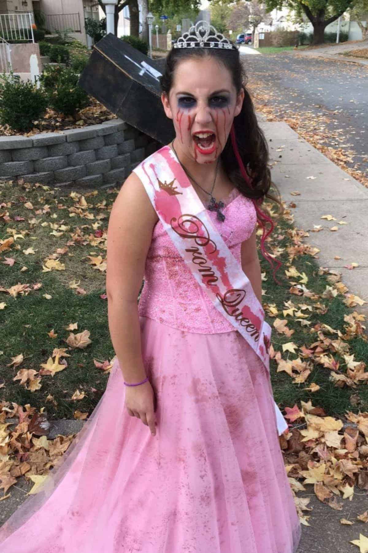 A girl in a pink prom dress and tiara with “Prom Queen” sash has fake blood on her face and mouth, standing outdoors with leaves on the ground and a coffin prop behind her.