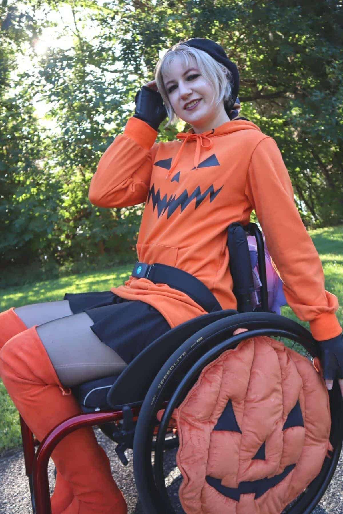 A person in a wheelchair dressed in a festive orange outfit with a jack-o’-lantern face sits outdoors, smiling at the camera.