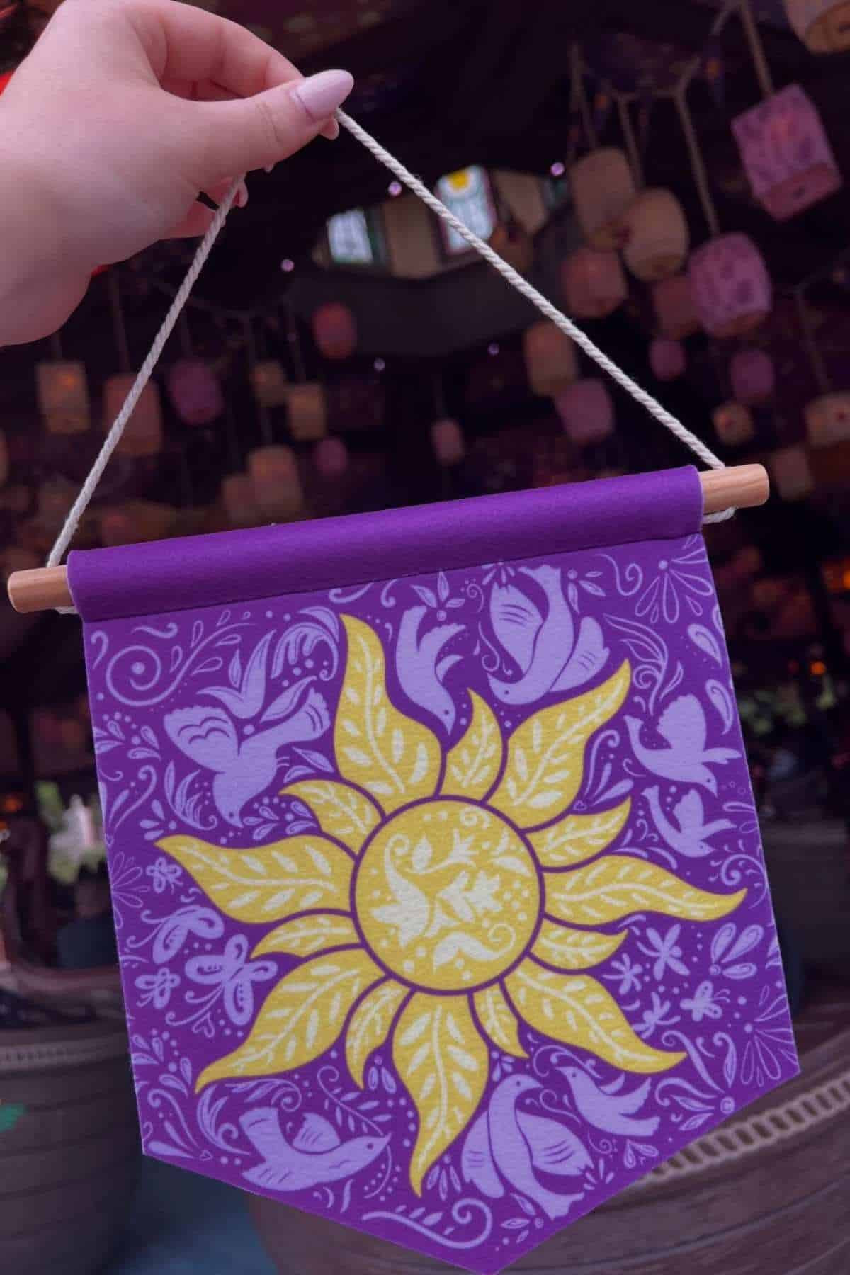 A hand holds a purple banner with a yellow sun and bird design, hung by a white string, with lanterns and dim lighting in the background.