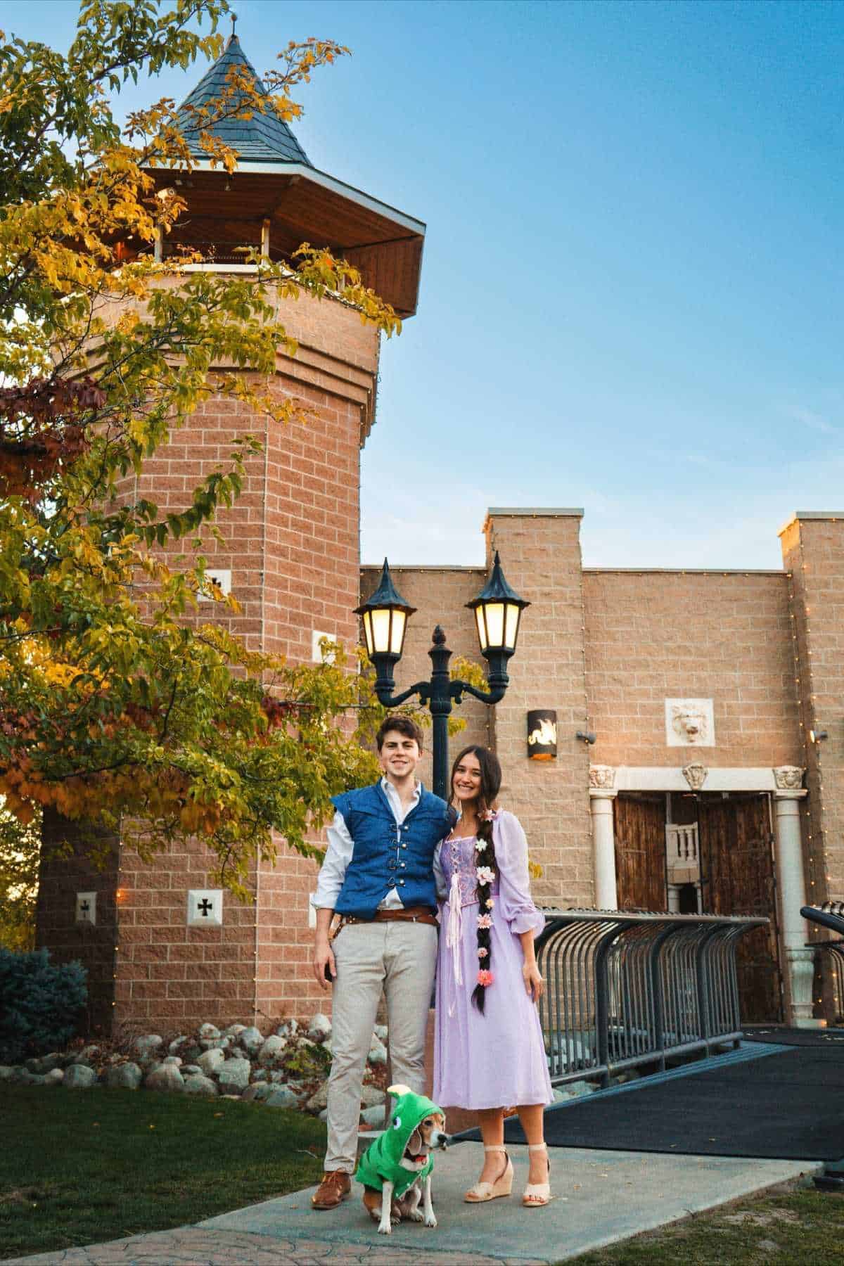 A man and woman stand smiling with a small dog by their side in front of a brick castle-like building and a lamppost, under a clear blue sky.
