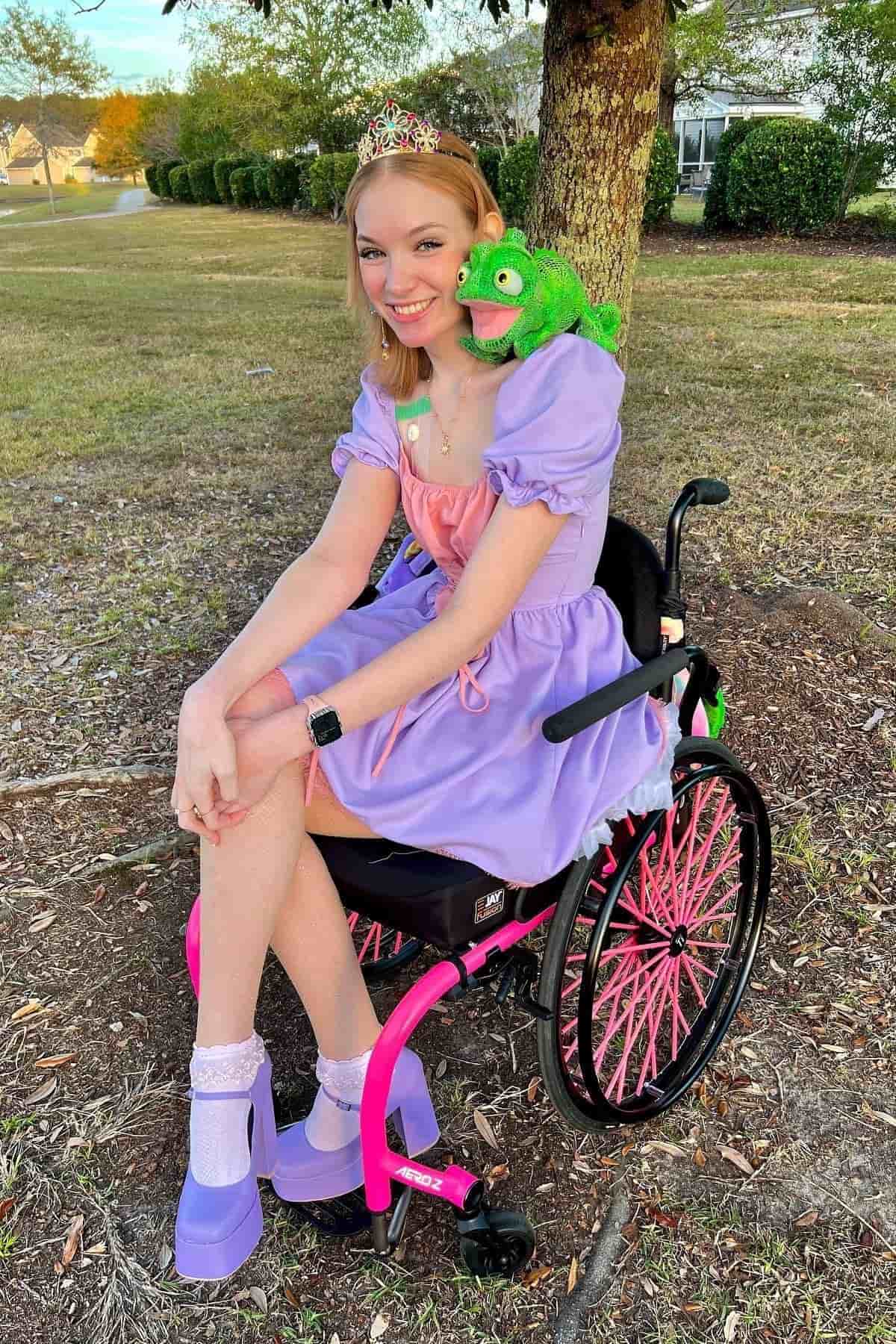 Woman in a wheelchair wearing a purple princess dress, tiara, and purple shoes, with a green plush chameleon on her shoulder, sitting outdoors near a tree.