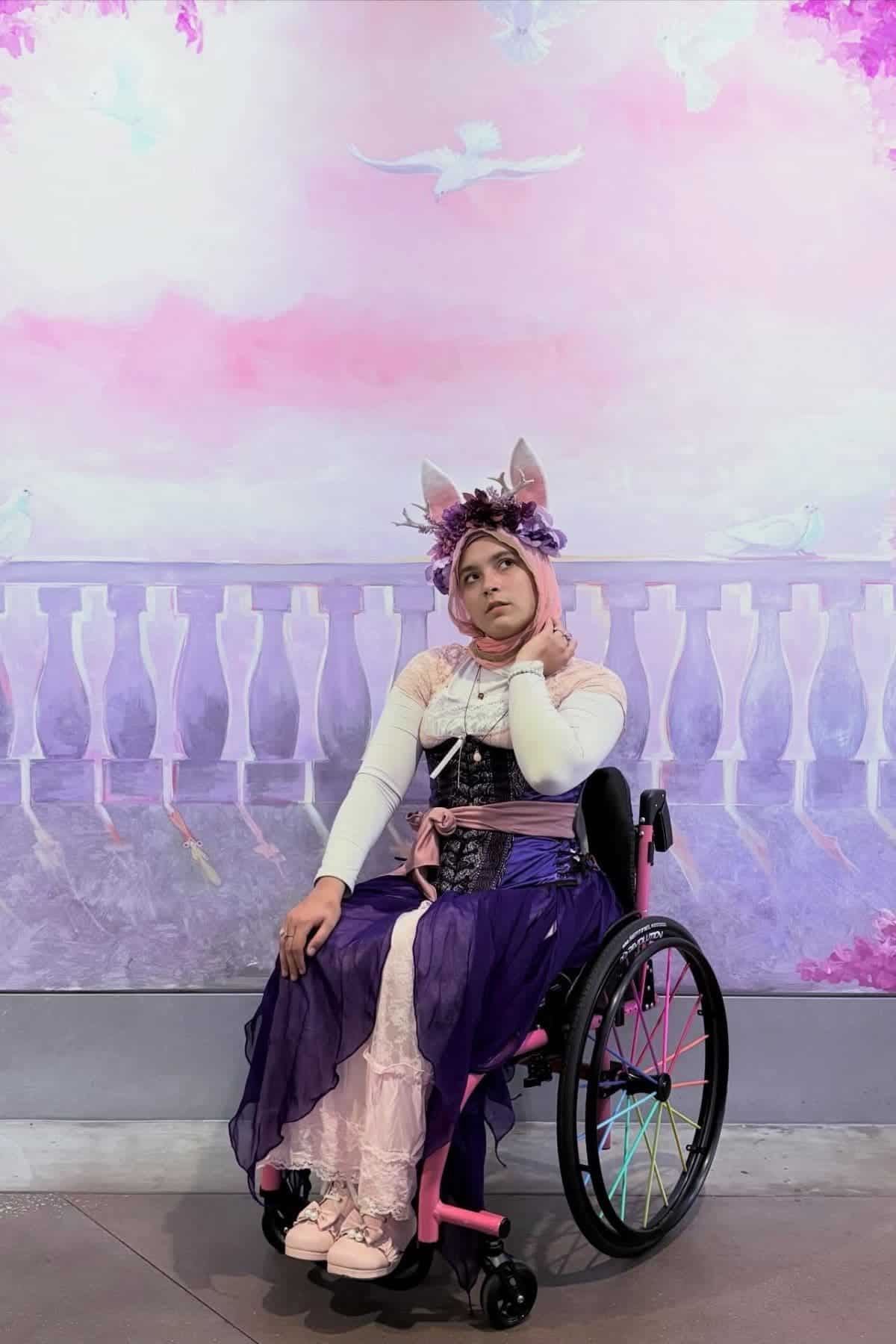 A person in a wheelchair wears a floral headpiece and layered pastel costume, sitting in front of a pink and purple painted backdrop with birds and a fence.
