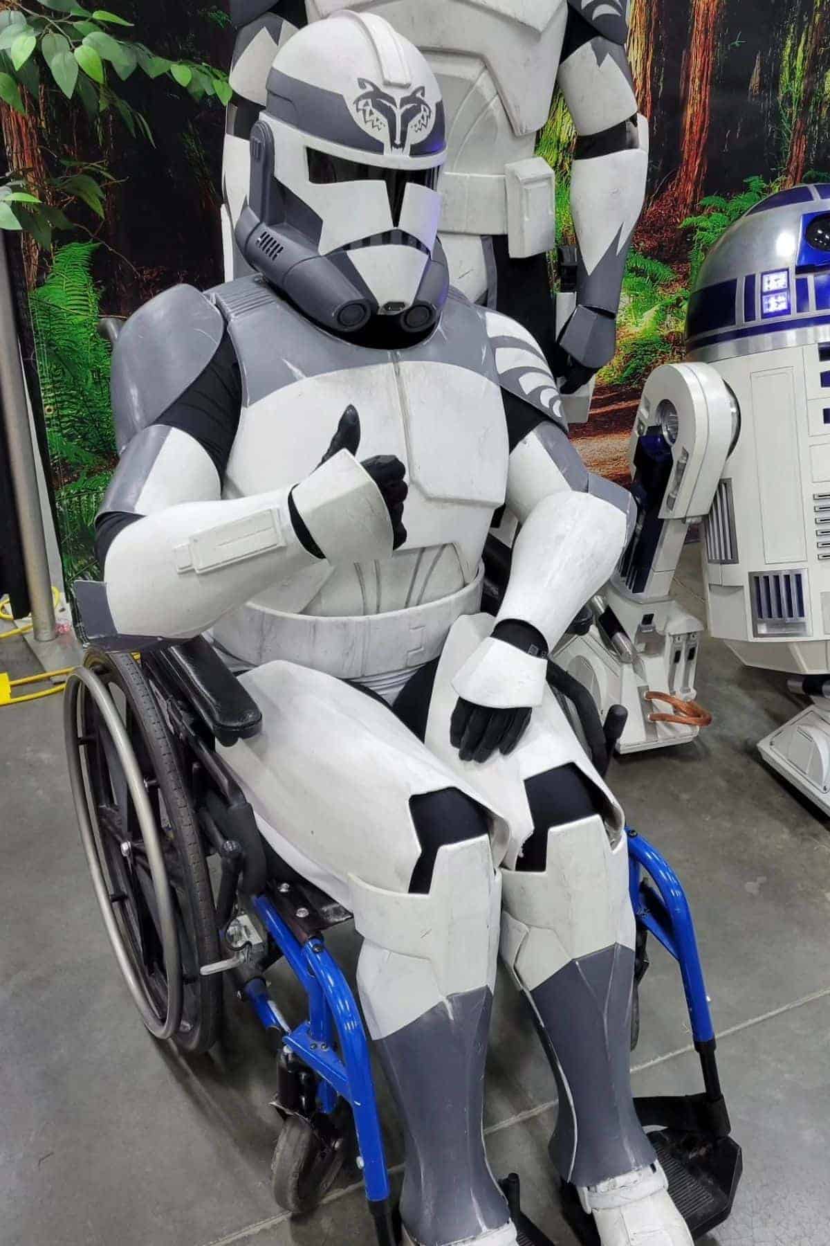 Person in a white and gray Star Wars clone trooper costume gives a thumbs up while seated in a blue wheelchair, with R2-D2 and another costumed figure in the background.